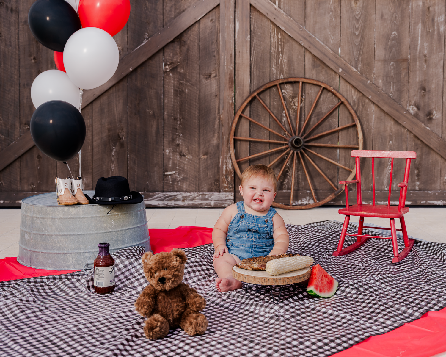 Baby and teddy bear sitting on checkered blanket with colorful balloons in rustic barn-style setting.