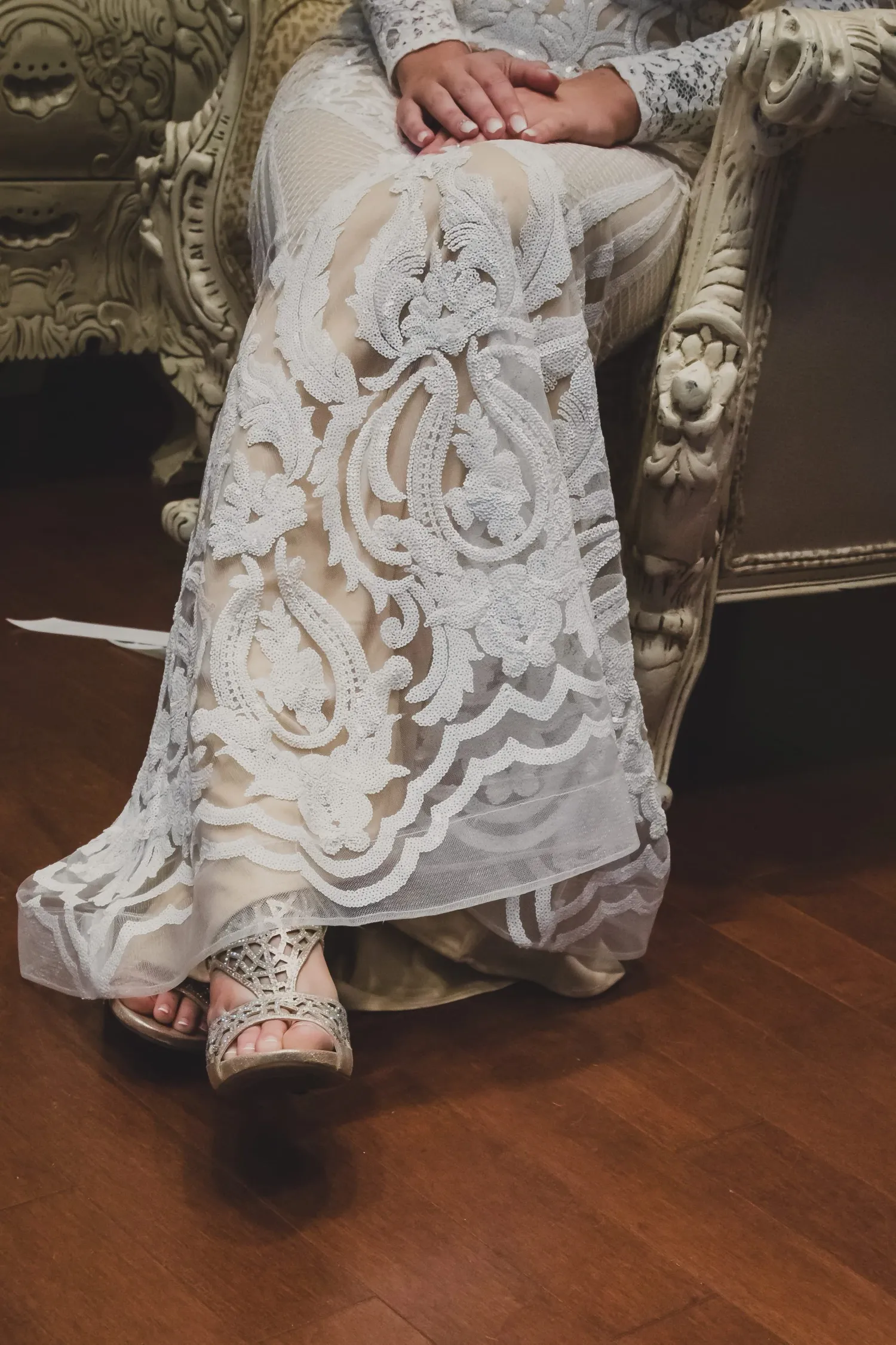 Intricate white lace wedding dress detail with ornate patterns shown against a vintage chair and wooden floor.