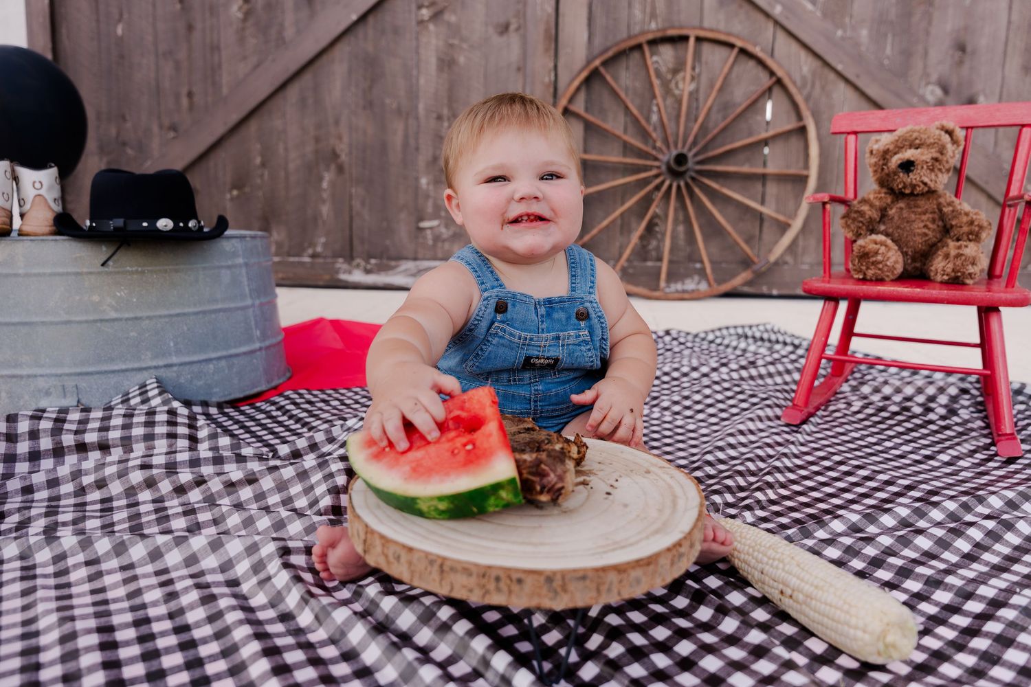 Baby in overalls sits on checkered blanket with watermelon slice during rustic outdoor picnic setting.
