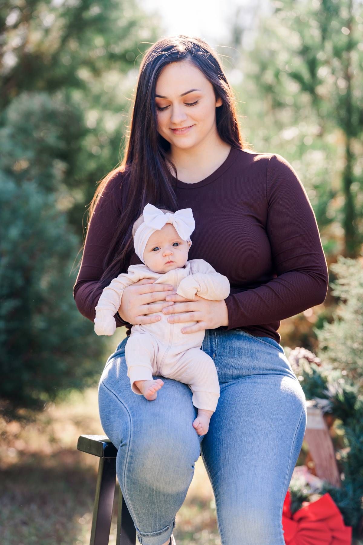 Mother in black shirt and jeans holds young baby wearing white bow on outdoor photoshoot.