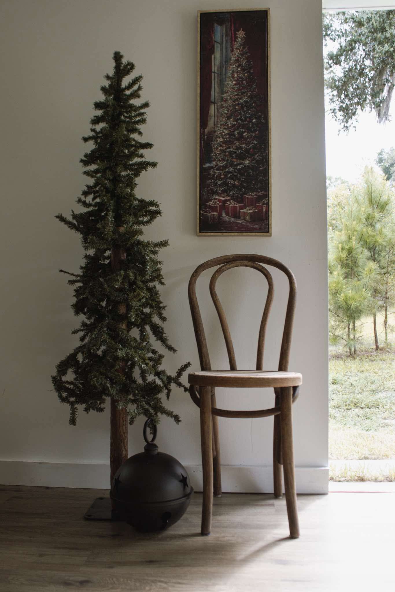 Rustic wooden Thonet chair beside a slim Christmas tree and festive wall art near a bright window.
