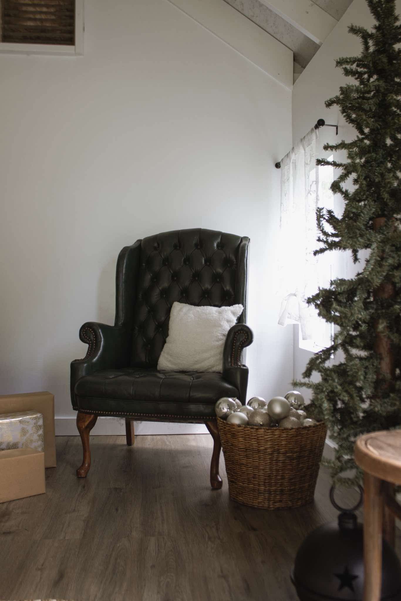 Dark leather tufted wingback chair with white pillow beside Christmas tree and wicker basket of ornaments in cozy room.