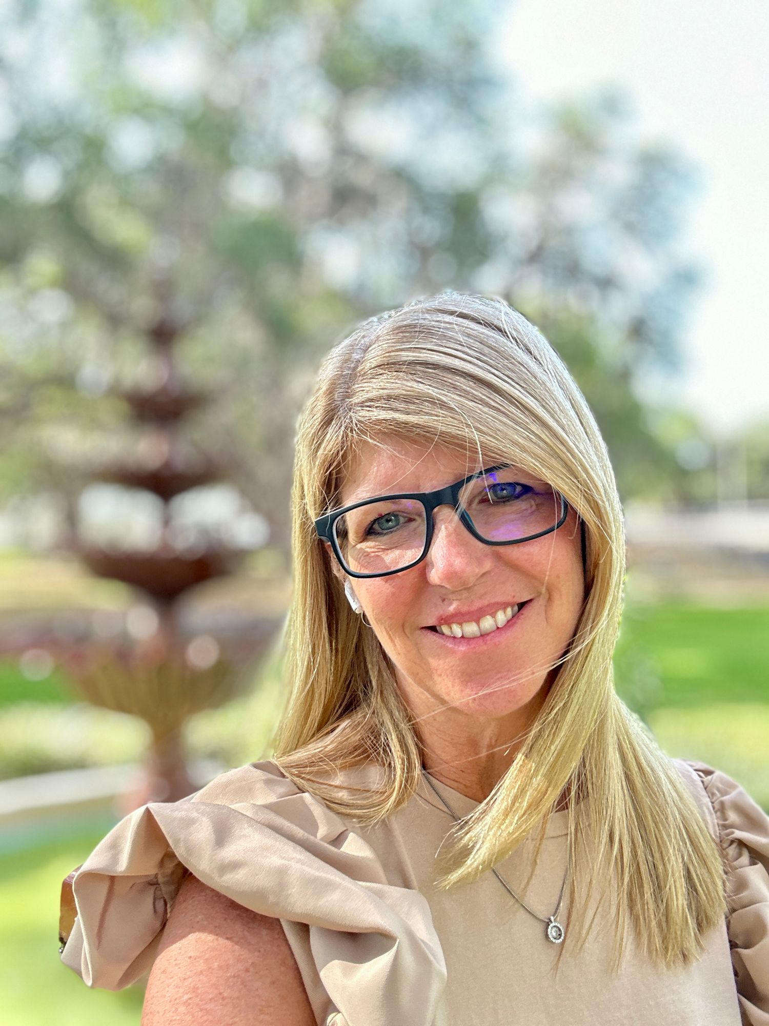 Portrait outdoors showing someone wearing glasses and a beige top with natural lighting and bokeh background.
