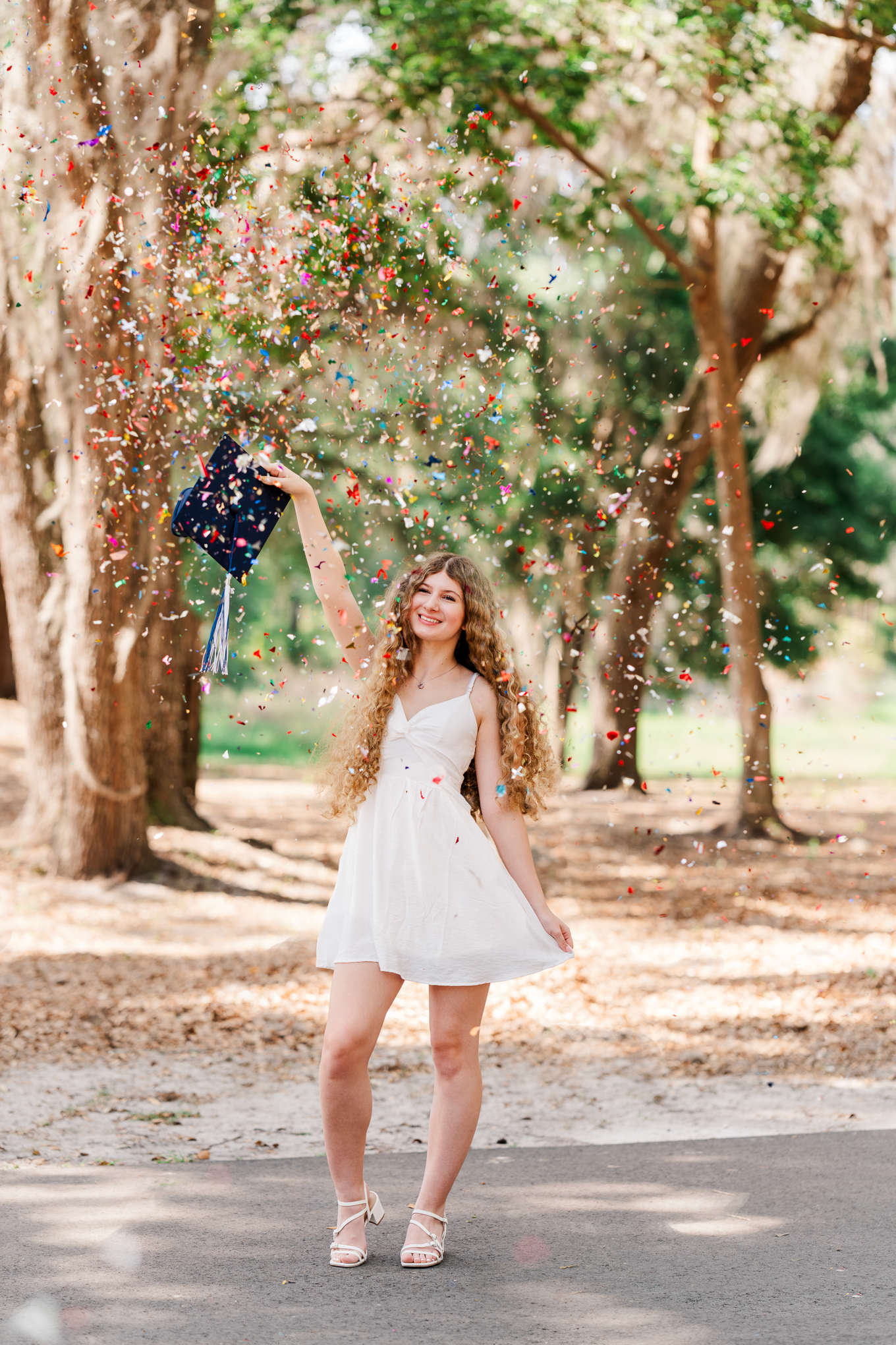 Graduate celebrates with confetti toss in white dress under tree canopy.