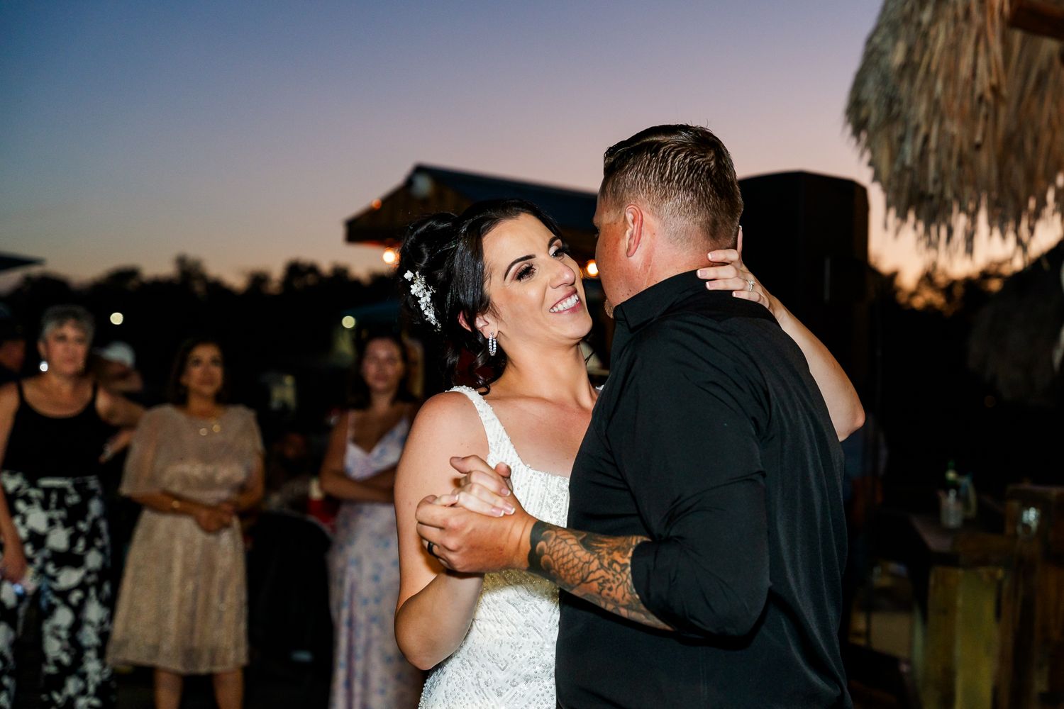 Two people slow dance together at an outdoor wedding reception during sunset.