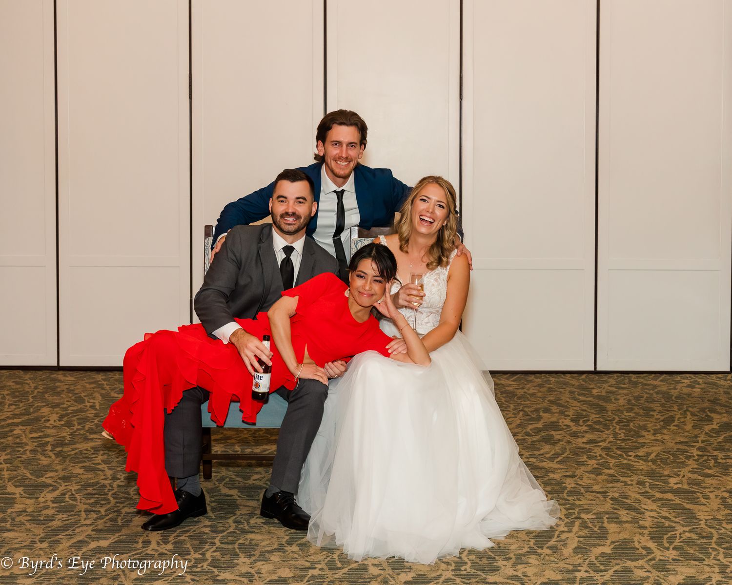 Wedding party poses playfully on beige carpeting against a cream colored wall.