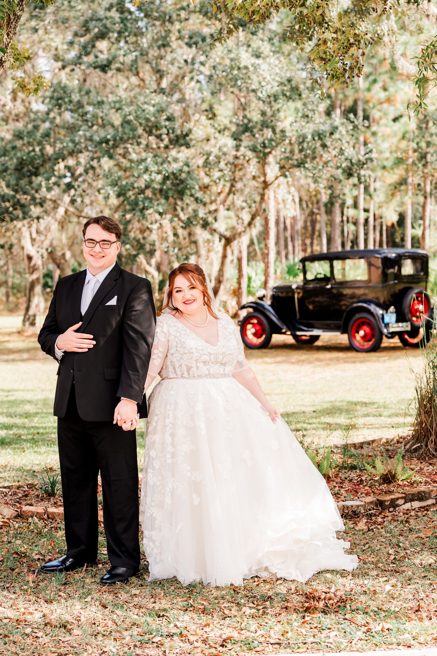 Wedding couple poses outdoors with vintage car against tree-lined backdrop.