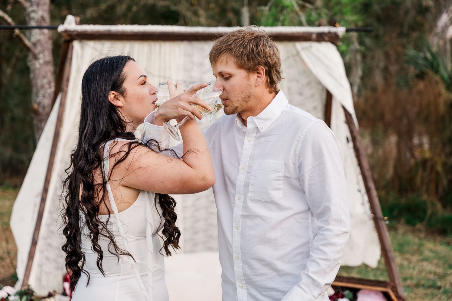 A couple shares a playful moment while feeding each other cake outdoors with a white tent in the background.