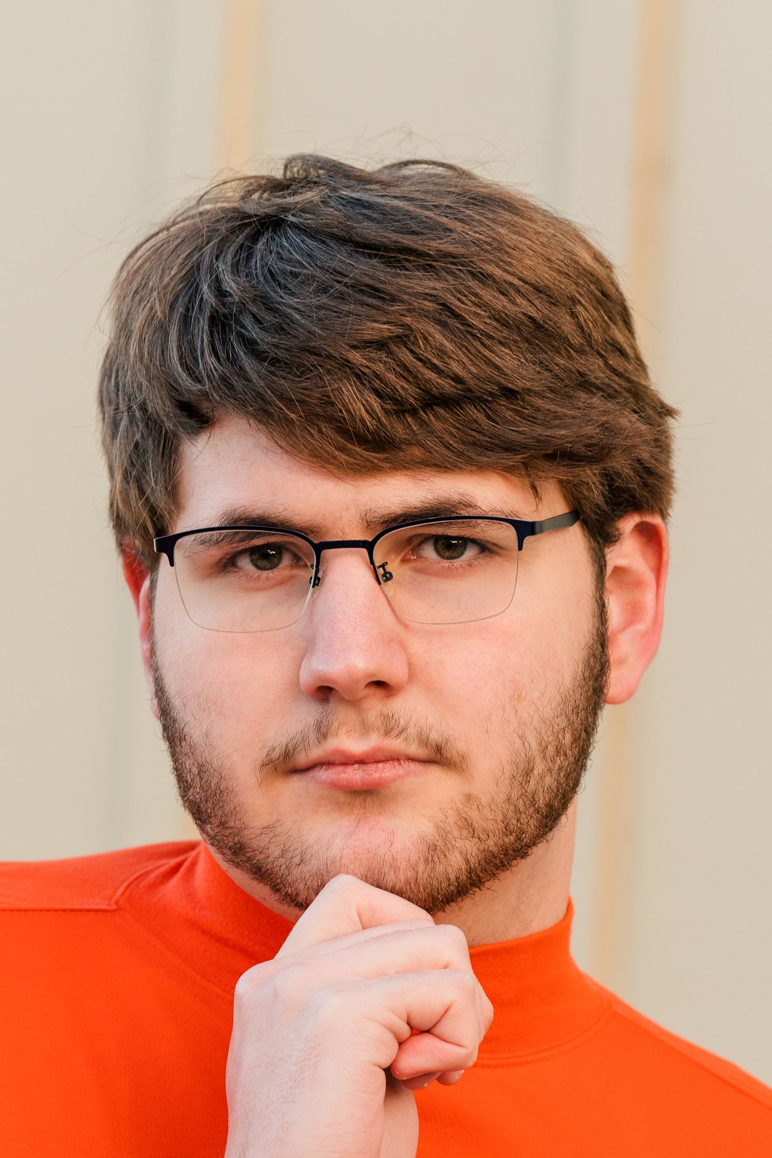 Portrait shot against beige wall wearing orange turtleneck and glasses.