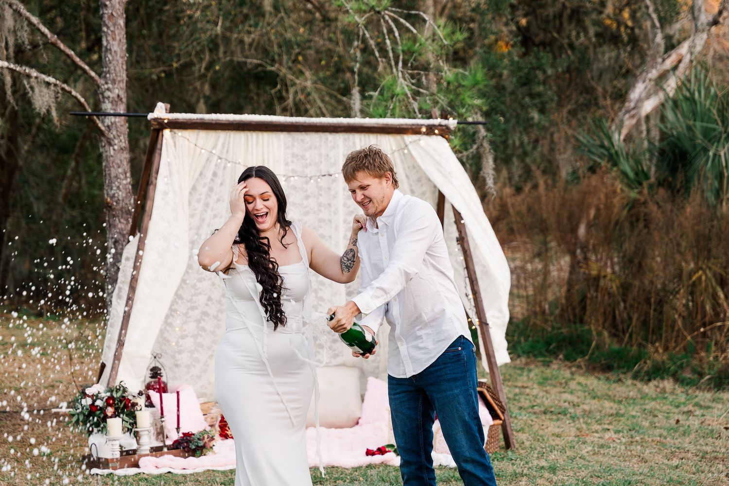 Couple in white outfits celebrating with champagne spray in an outdoor garden setting.