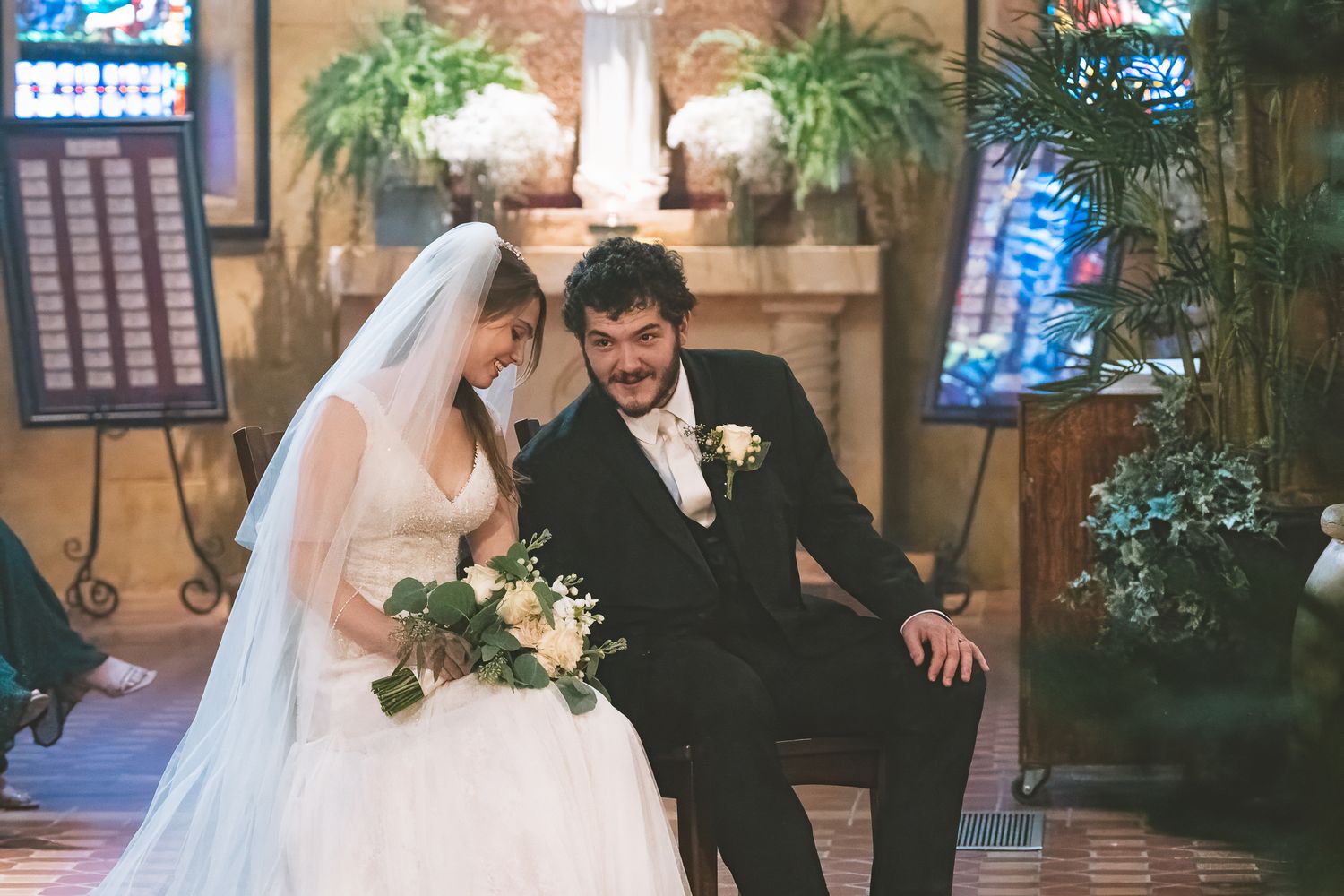 Wedding ceremony in a chapel with blue stained glass windows and indoor plants.