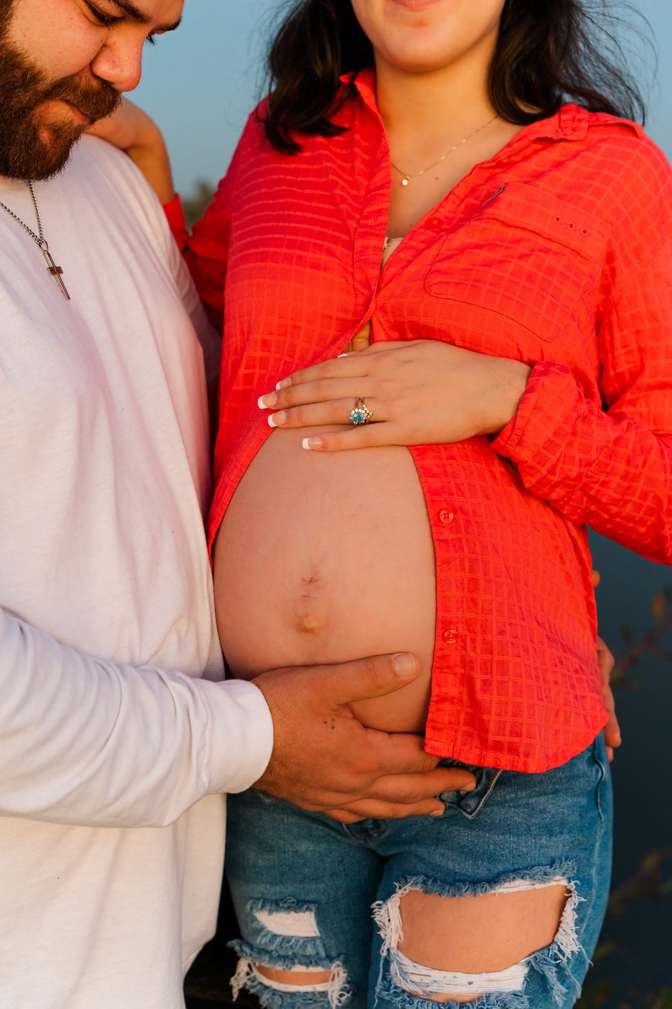 A couple poses together, with the woman wearing a red top and ripped jeans in a maternity photo shoot series.