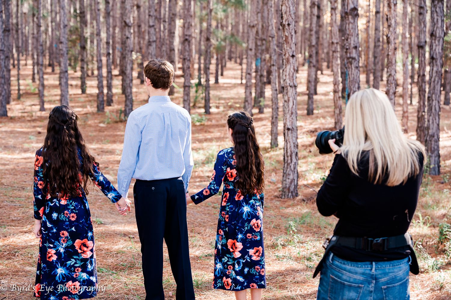 Behind the scenes of Kathy photography siblings walking together through a pine forest on a sunny afternoon.