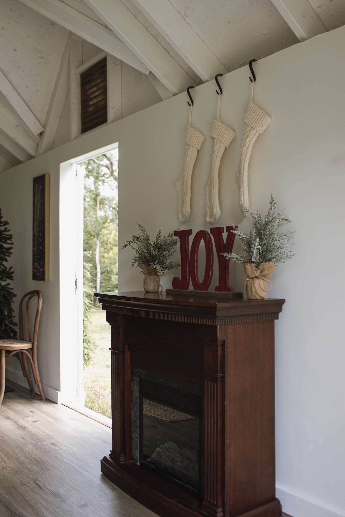Rustic fireplace mantel with red JOY letters, Christmas stockings, and greenery in a cozy white shiplap room.