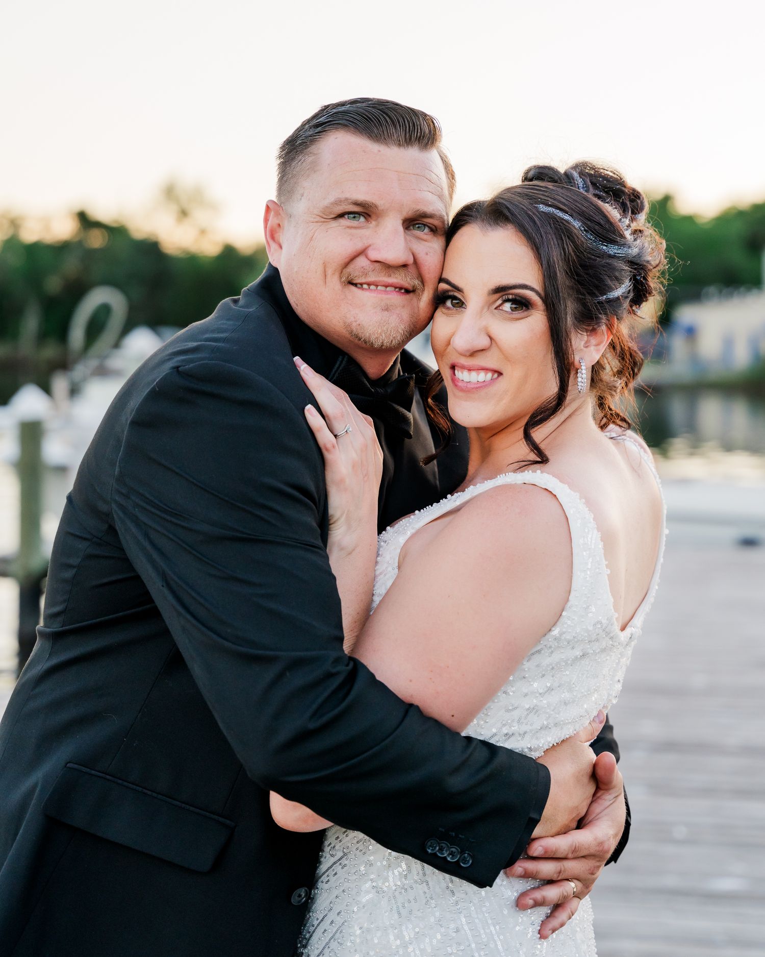 A couple in wedding attire embrace outdoors during sunset, with the bride in white and groom in black formalwear.