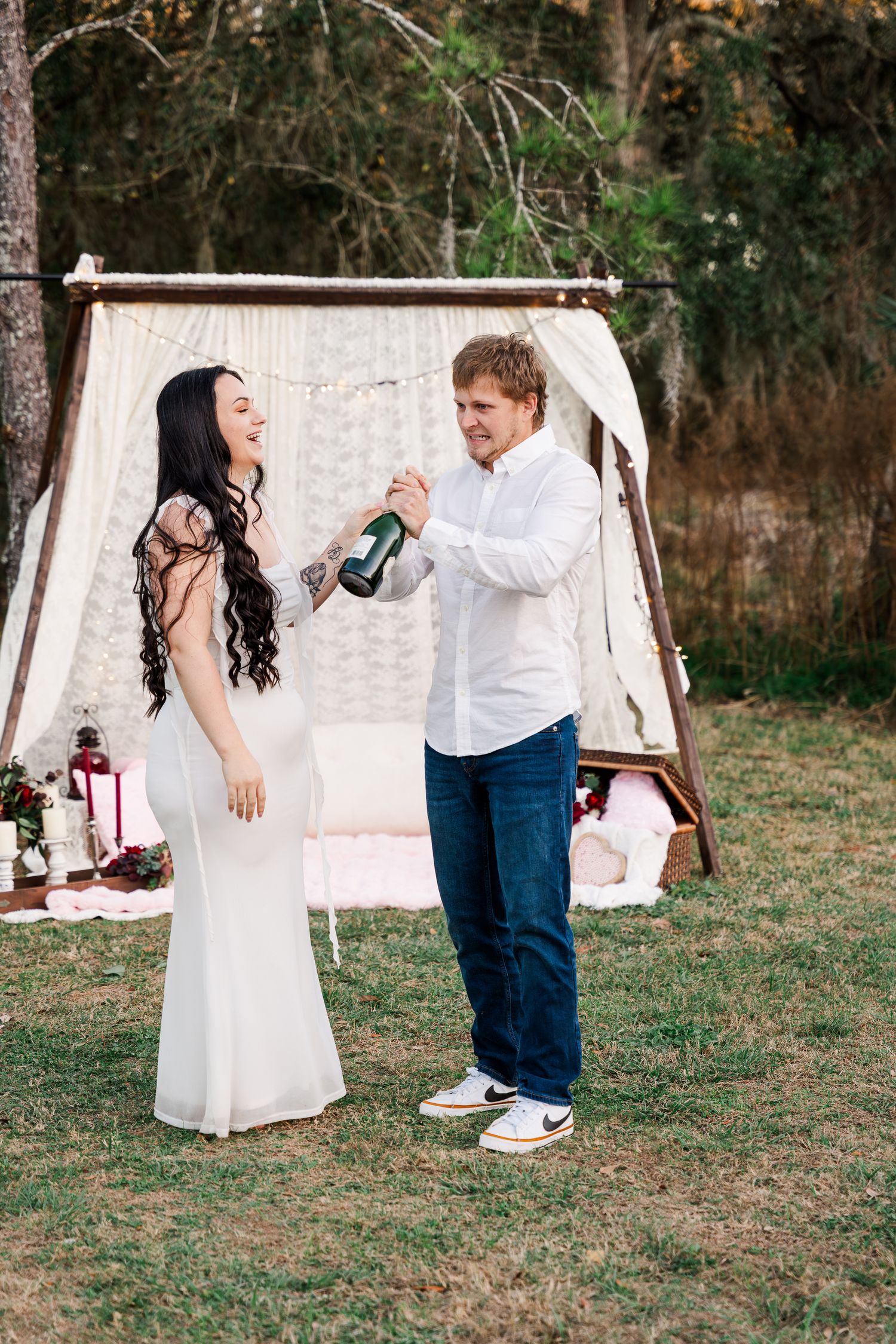 A couple celebrates with champagne in front of a white backdrop tent during an outdoor celebration.