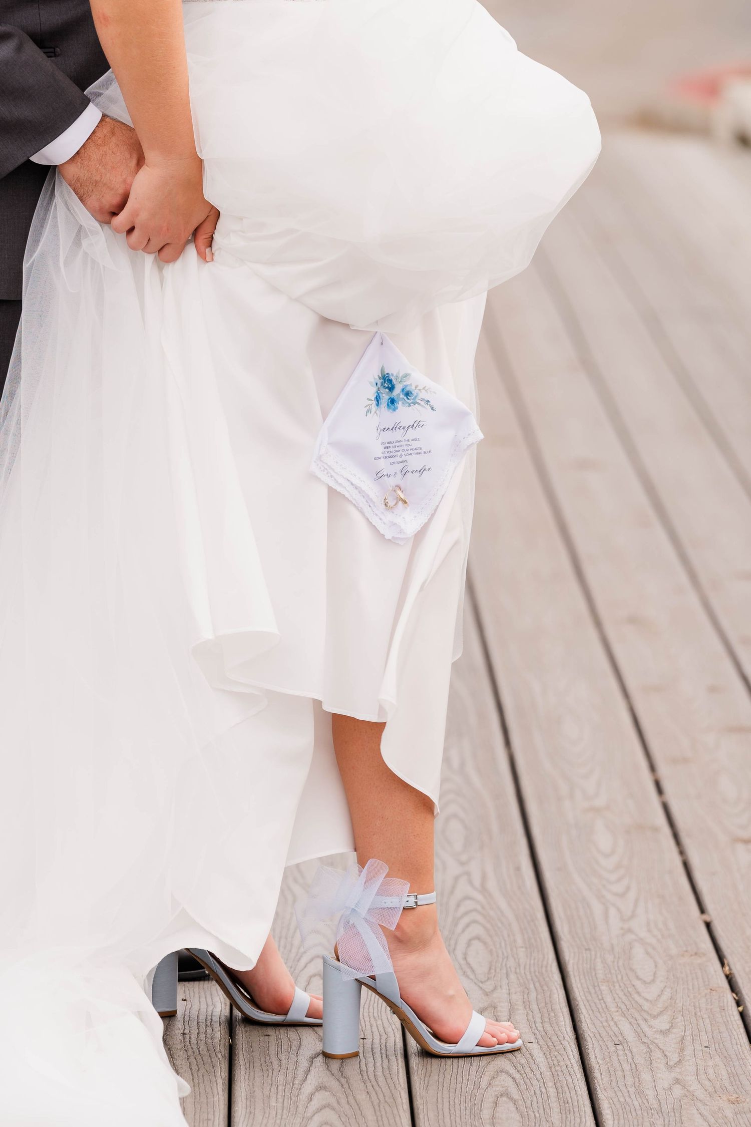 Close-up view of a bride's feet in clear heels and a white wedding gown on a wooden deck.