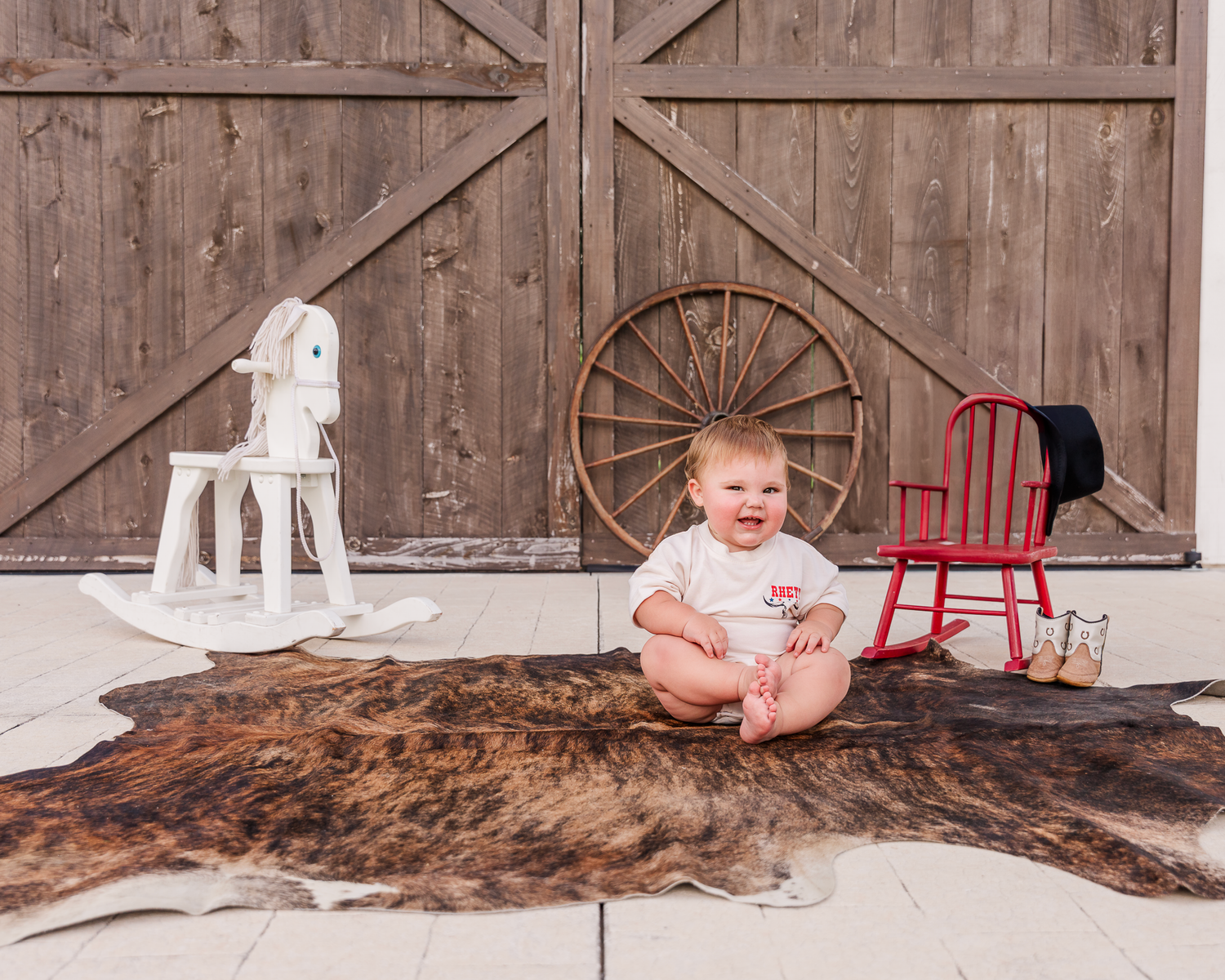 Small child sits on a brown animal hide rug with rustic barn doors and vintage rocking horses in the background.