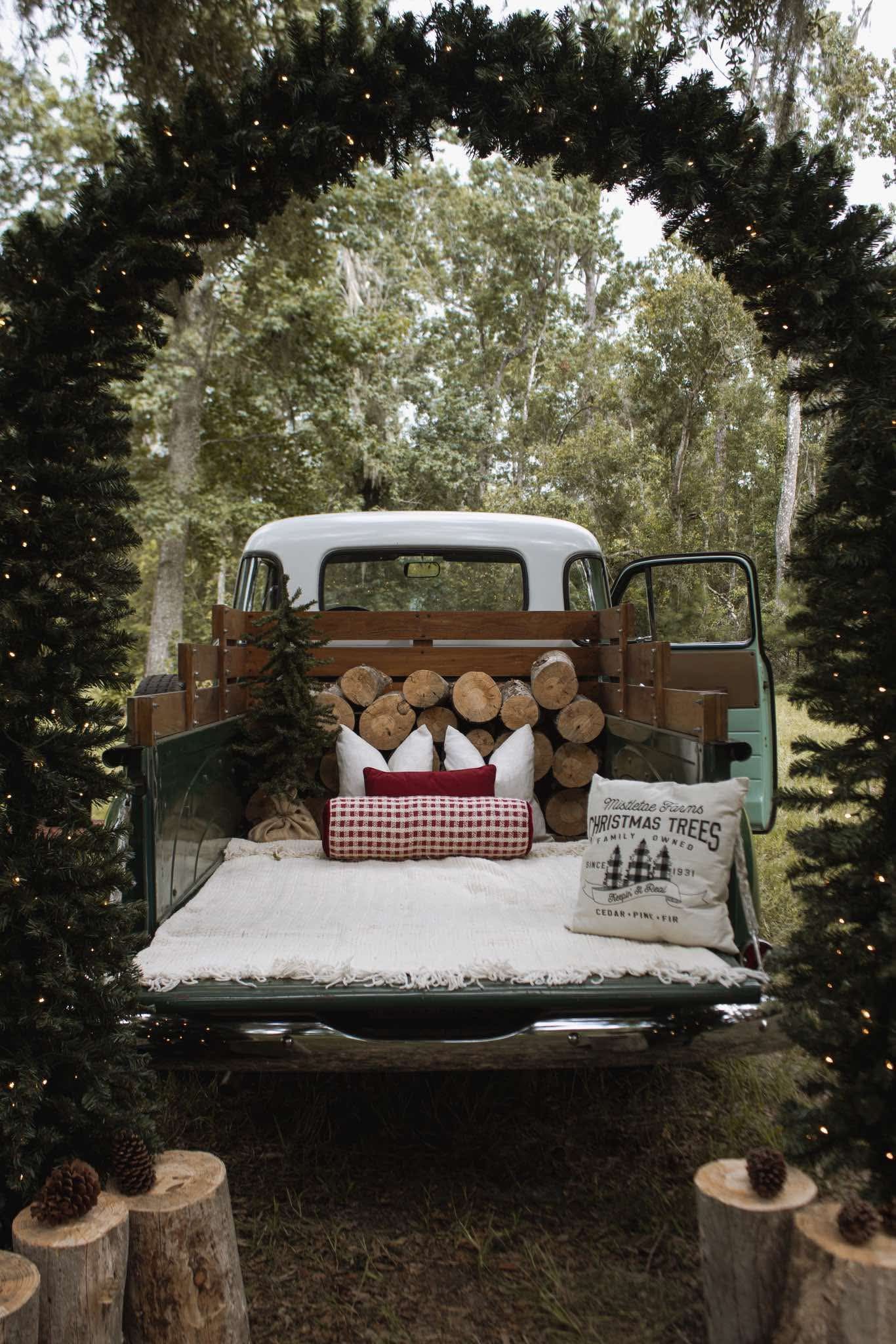 Vintage truck bed decorated with logs, plaid pillows, and Christmas tree signs under a festive evergreen arch in a forest.