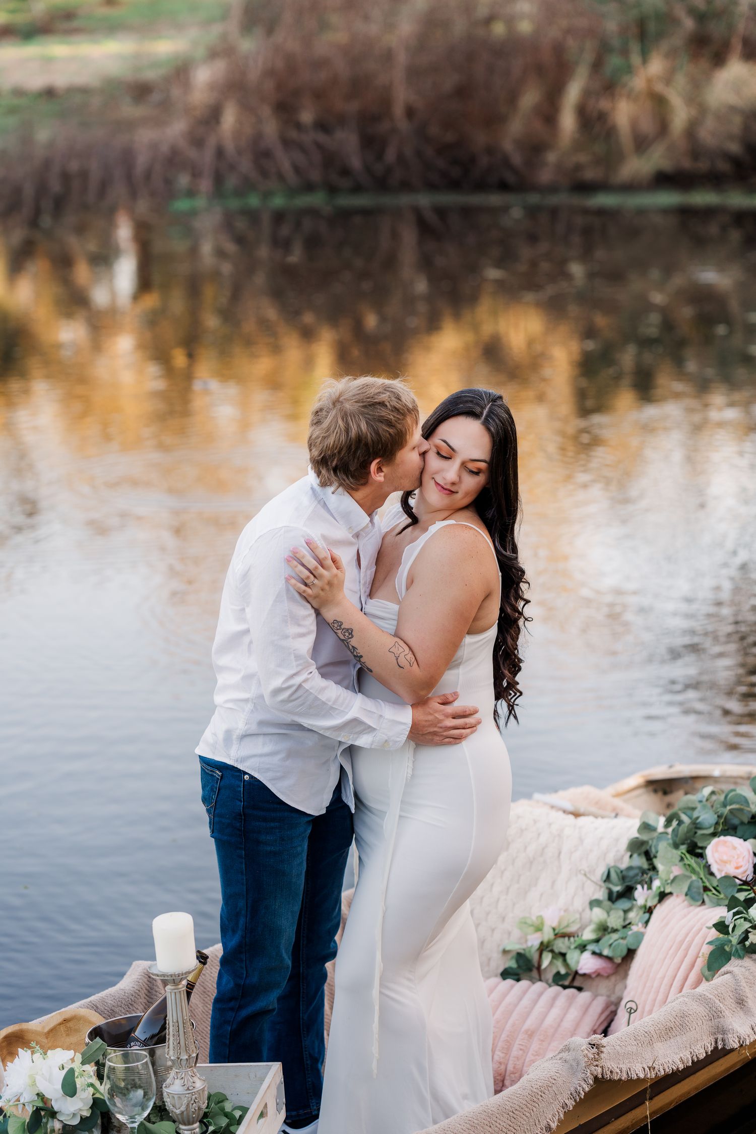 A romantic lakeside photoshoot with floral arrangements and candles on a wooden dock at sunset.