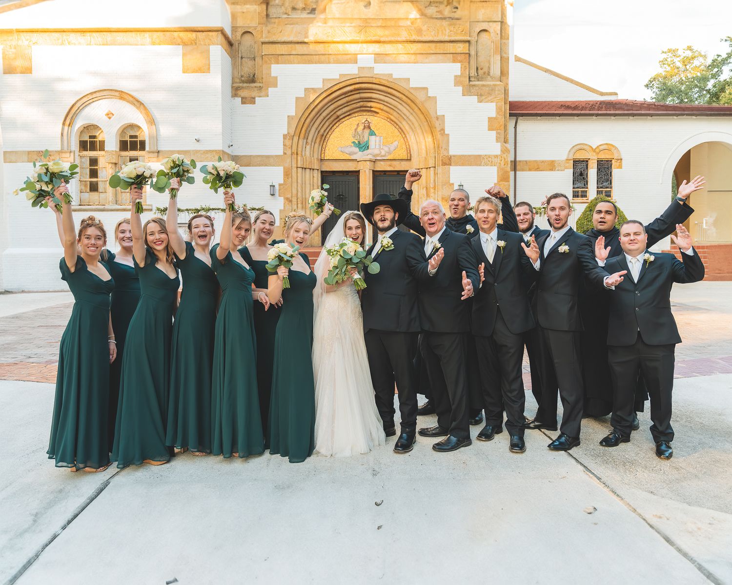 Wedding party in emerald green dresses and black suits posed outside a church building.