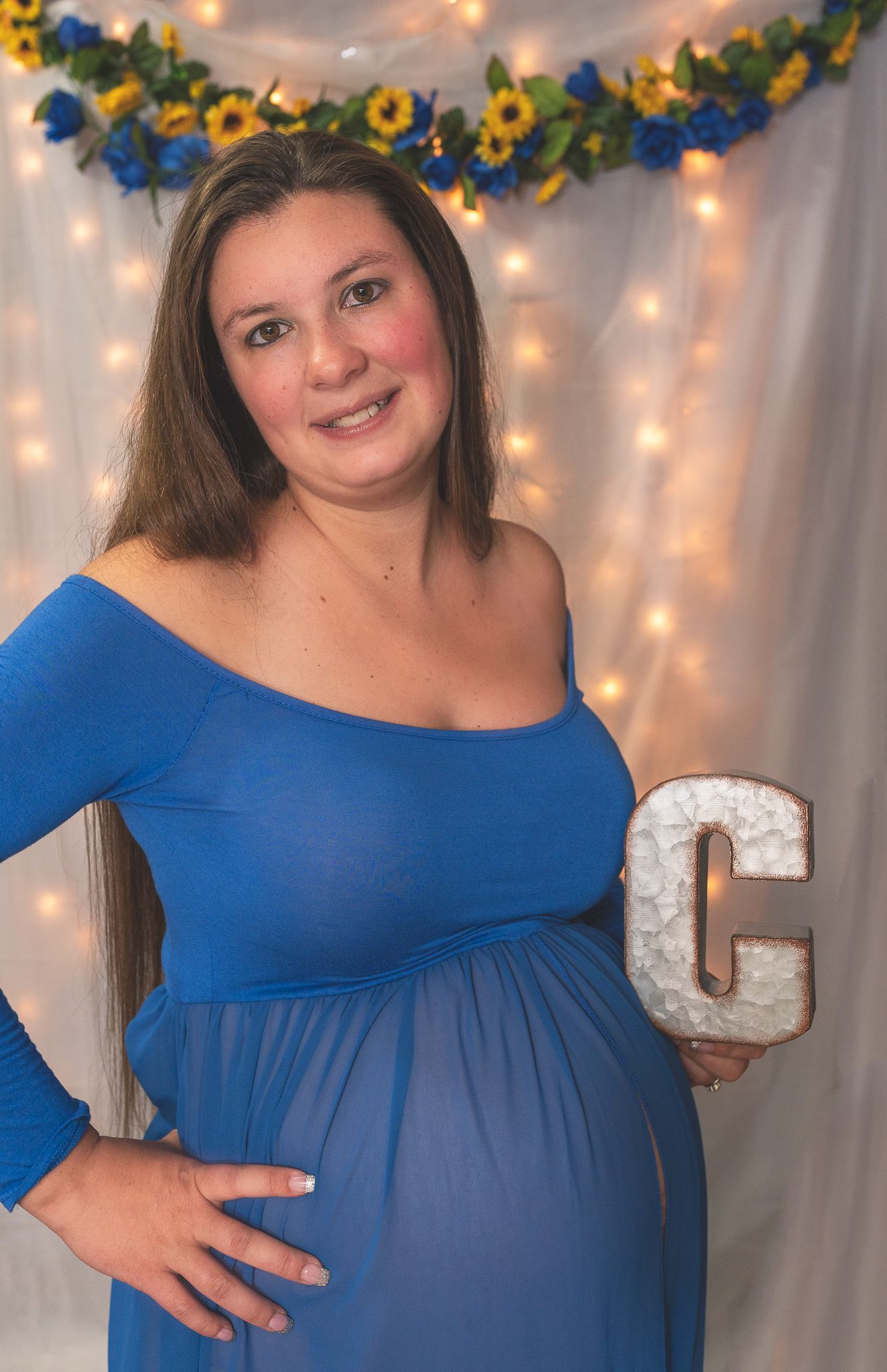 A pregnant woman in a royal blue maternity dress poses against a backdrop of sunflowers and string lights.