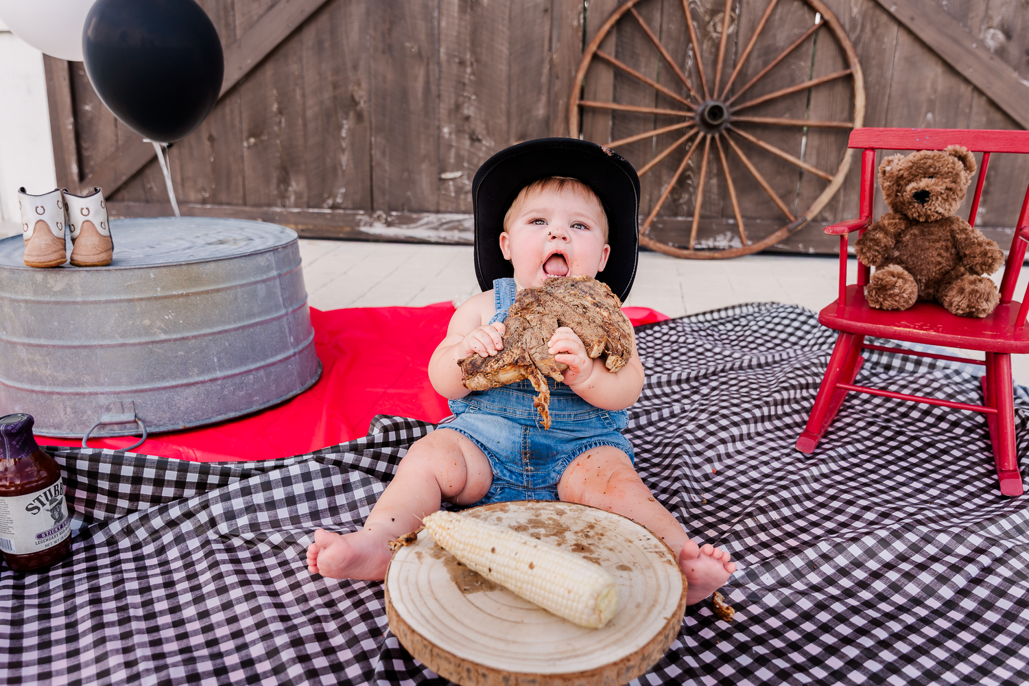 Small child on checkered blanket holding teddy bear during rustic themed photo shoot with vintage props.