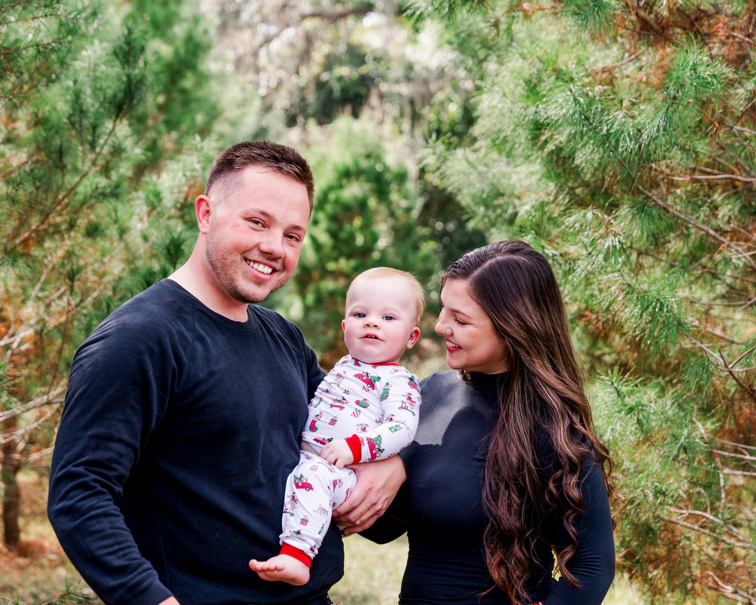 A smiling family of three poses outdoors among pine trees, with parents in black holding a happy baby in festive pajamas.