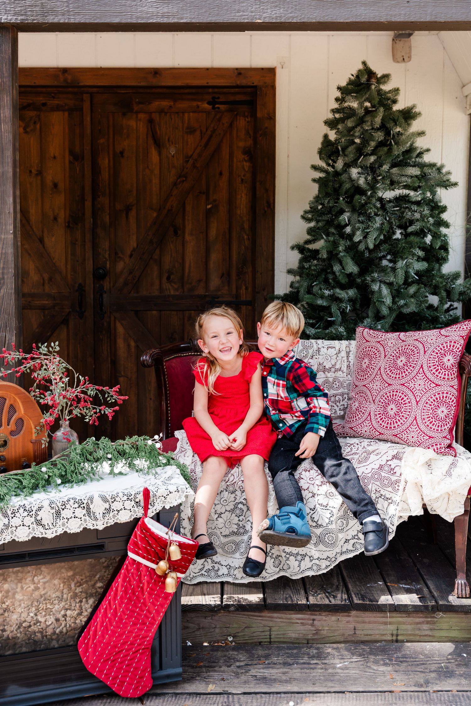 Two children in festive attire sit on a porch bench with Christmas tree and holiday decorations.