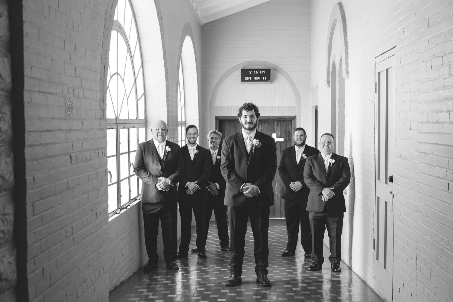Wedding party in formal attire posed along a white hallway with natural window lighting.