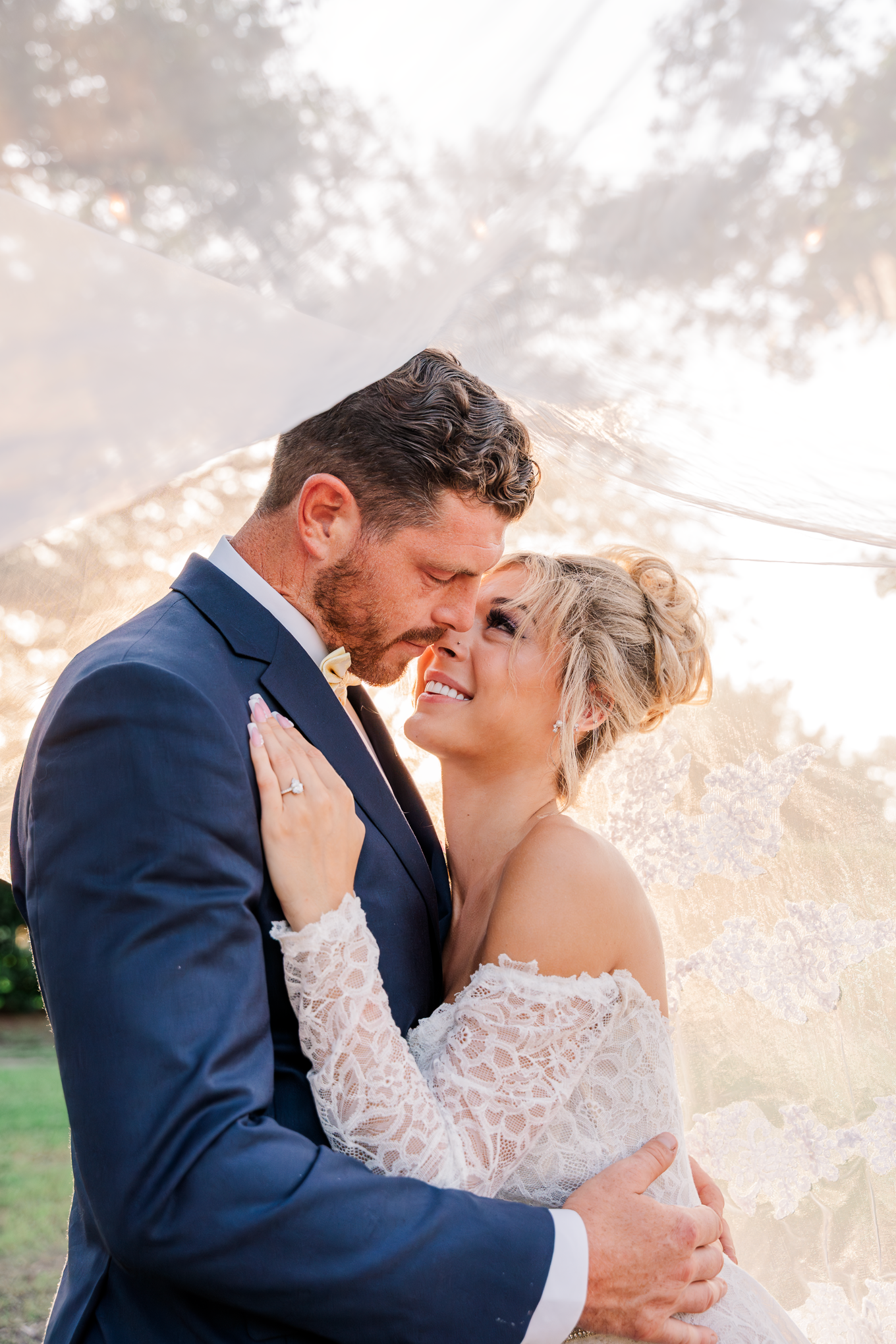 A romantic wedding couple embraces during golden hour as sunlight filters through lace while sharing an intimate moment.