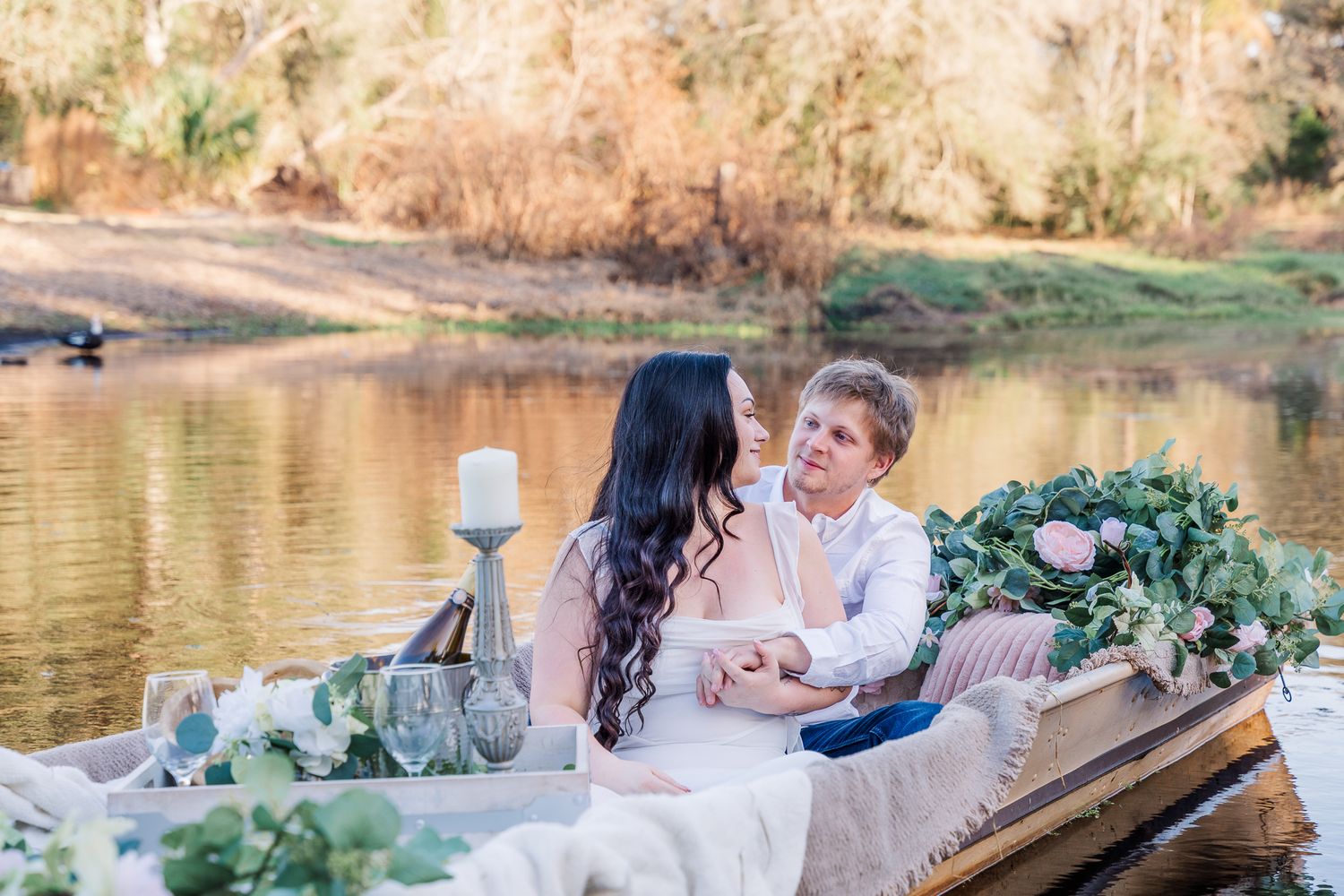A couple sits in a rustic wooden canoe adorned with floral decorations on a serene lake during  their sunset engagement session.