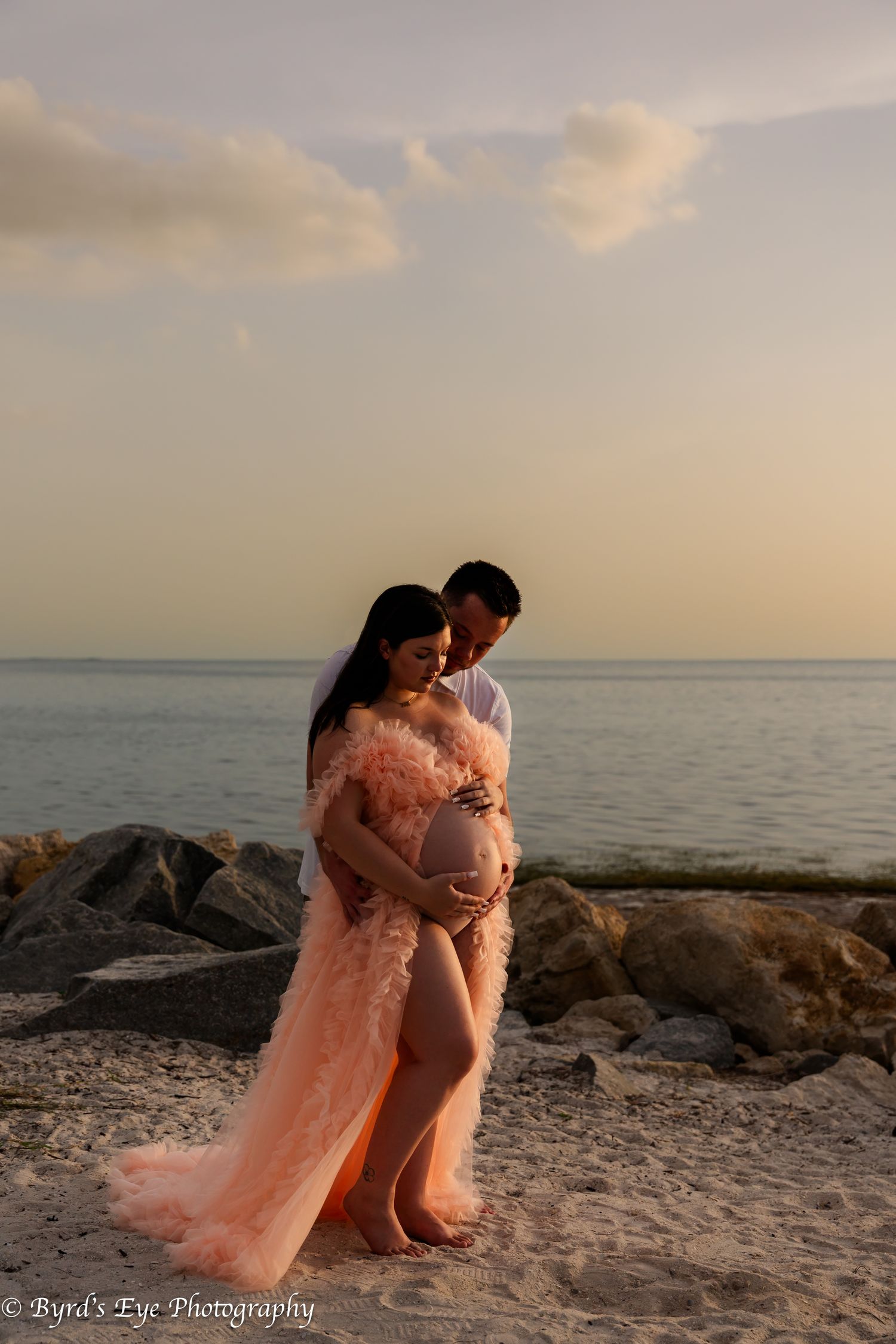 Romantic beach maternity photoshoot at sunset with couple embracing on rocky shoreline in soft peach gown.
