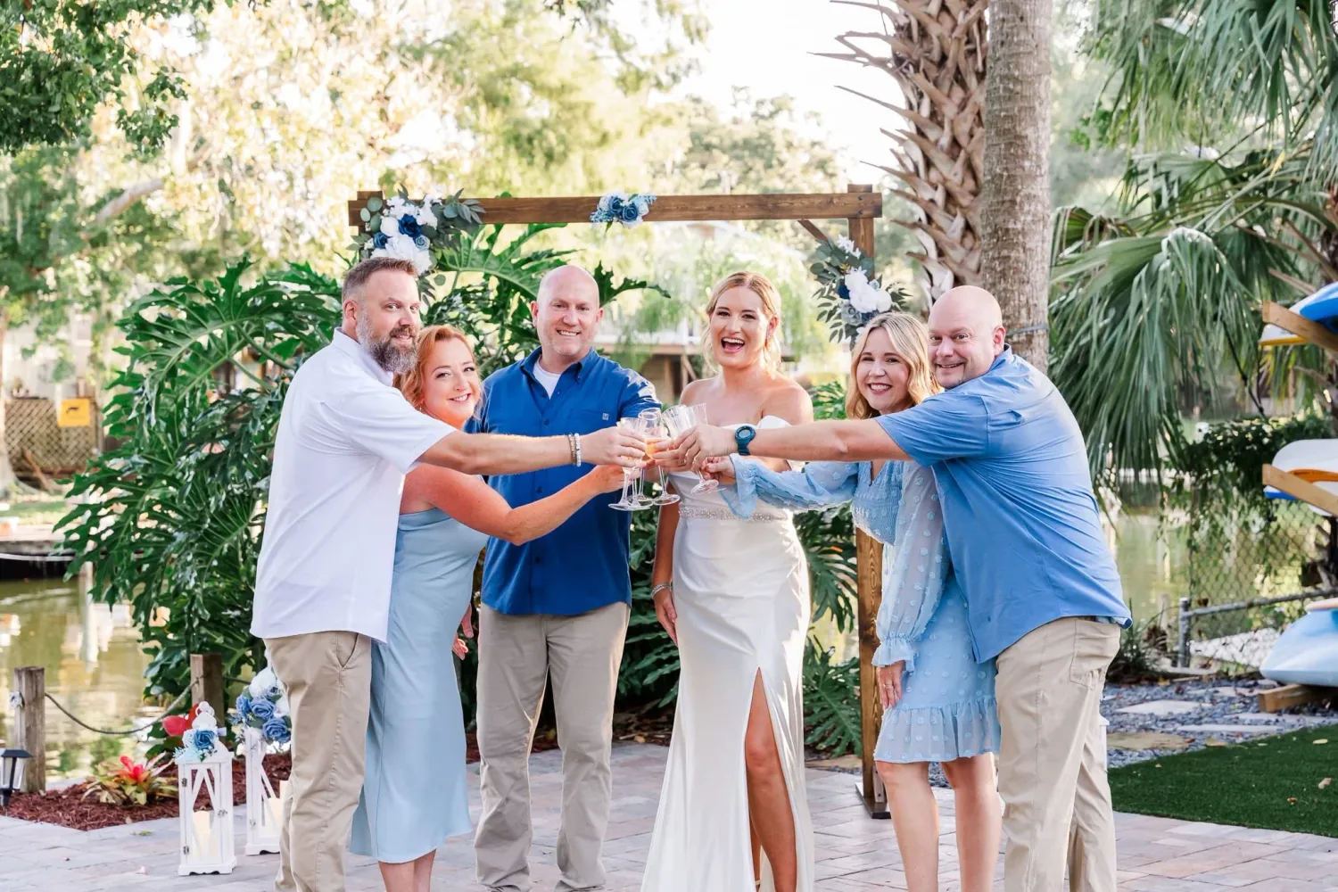 A group toasts with beverages outdoors in a tropical garden setting with palm trees and lush greenery.
