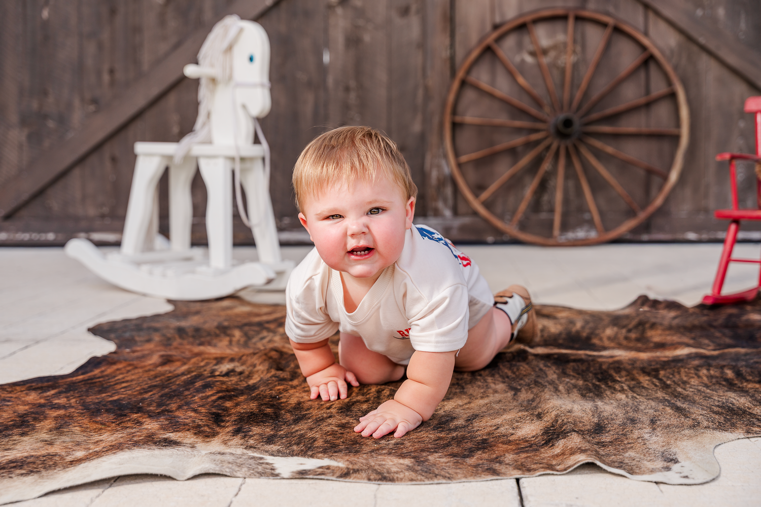Baby in white shirt lies on fur blanket with wooden rocking horse and wagon wheel decor in rustic setting.