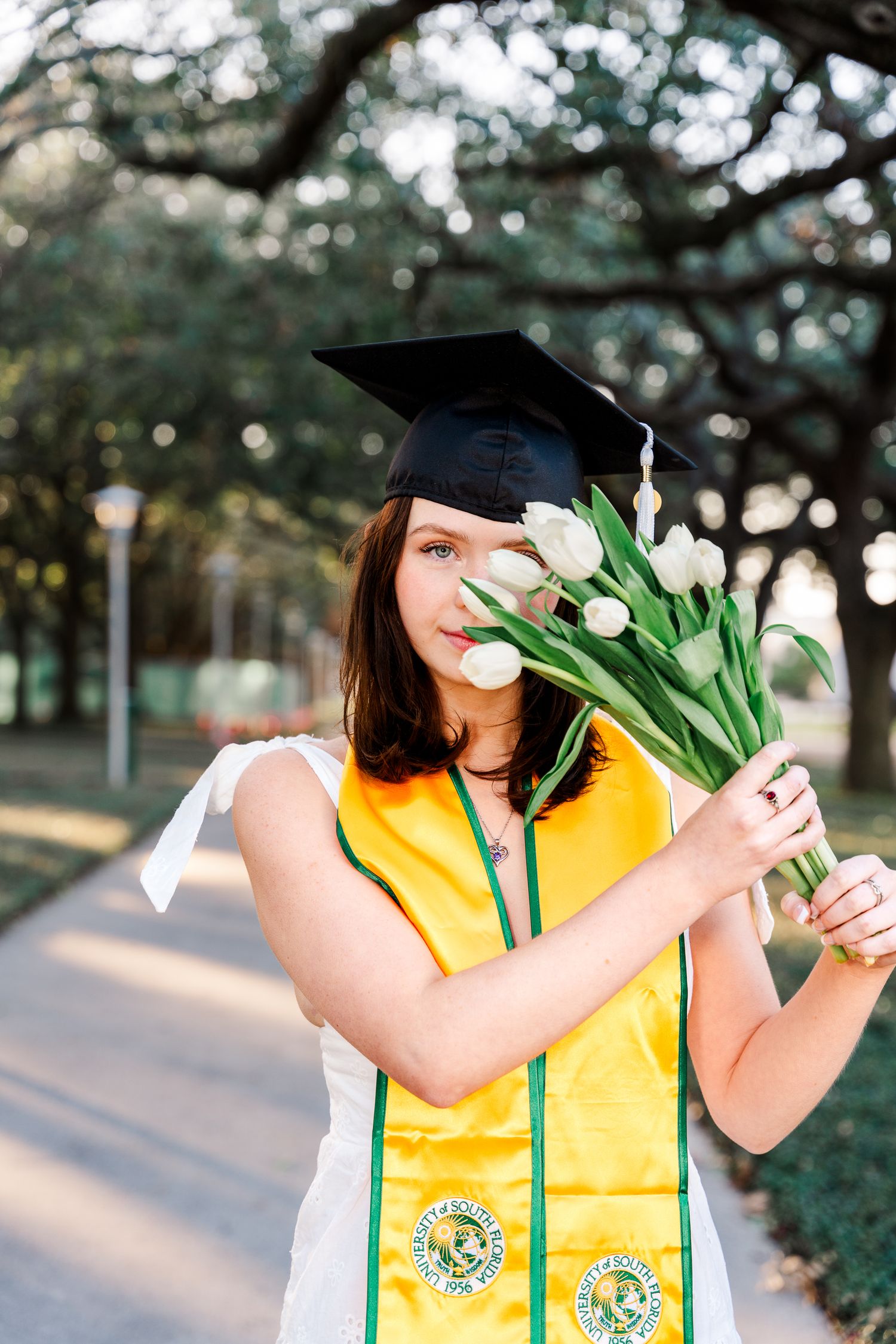 University of South Florida Graduate holding white tulips against a tree-lined backdrop during outdoor graduation photoshoot.