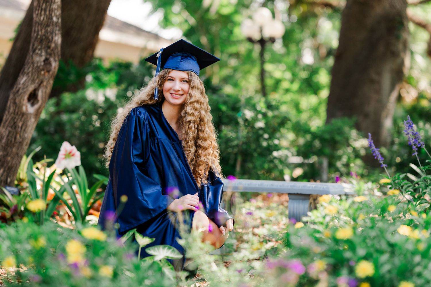 Graduate in blue cap and gown poses in garden setting with blooming flowers.
