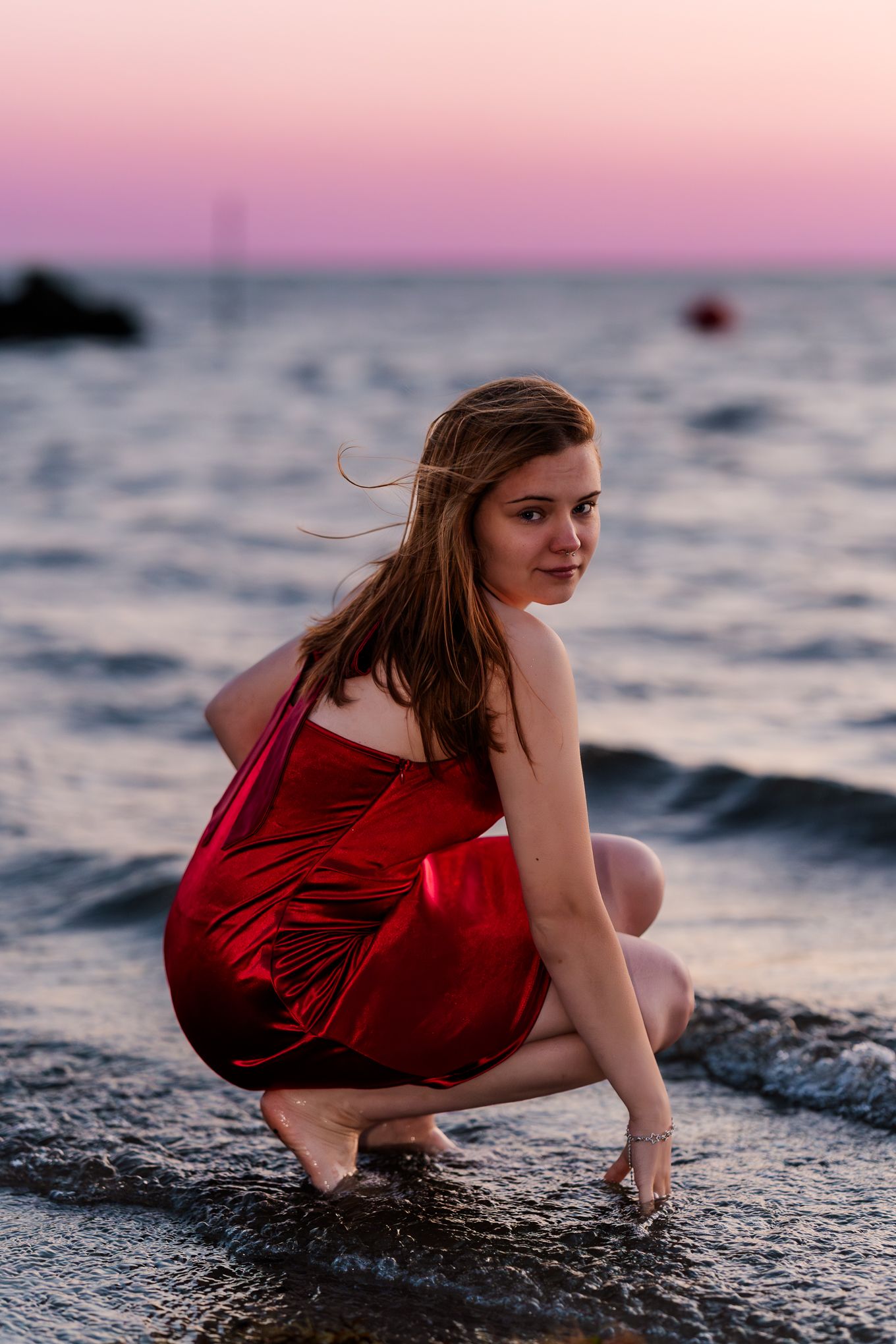 Figure in red dress poses dramatically on rocks by ocean at sunset.