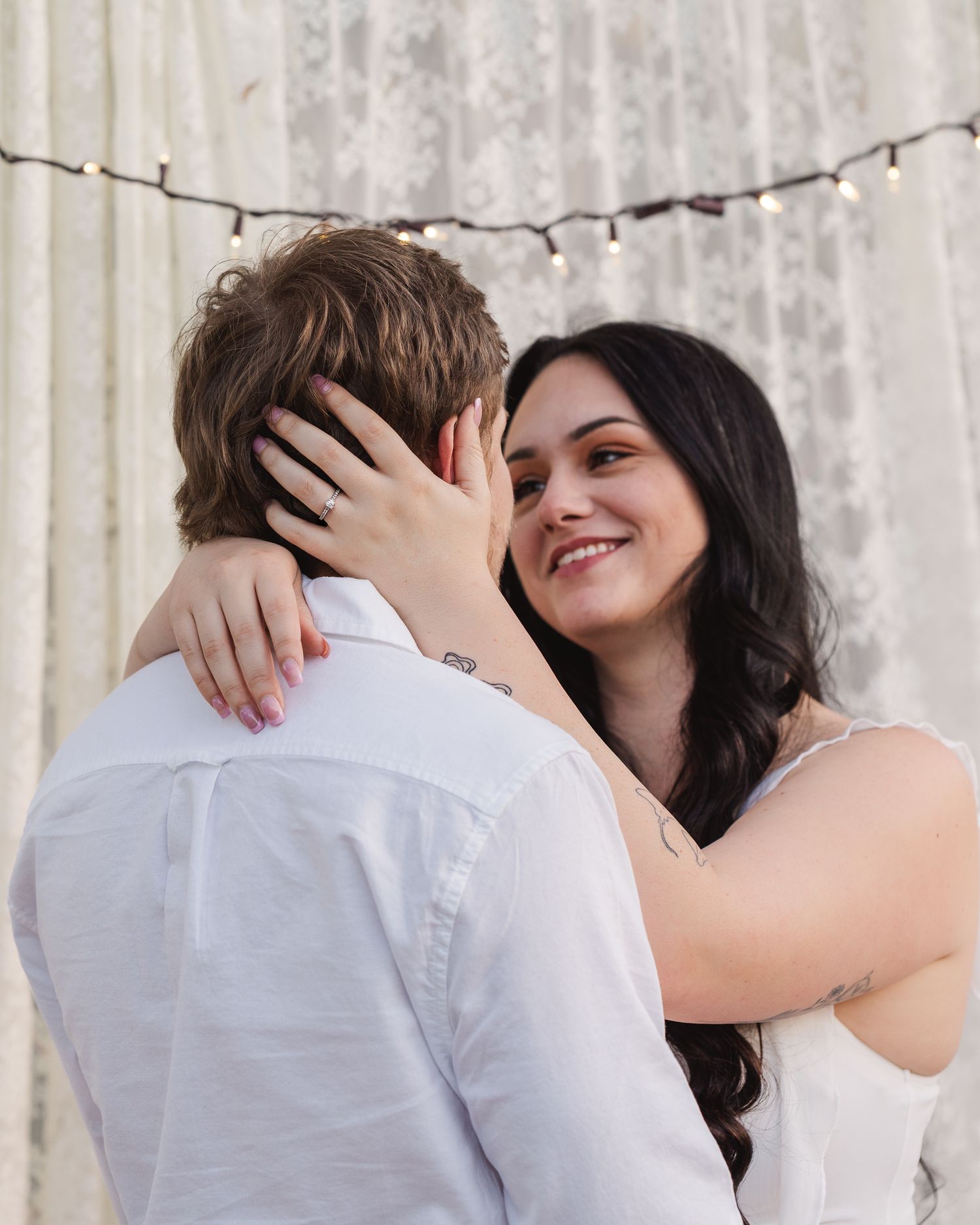 Couple in romantic embrace sharing tender kisses against white curtain backdrop with string lights.