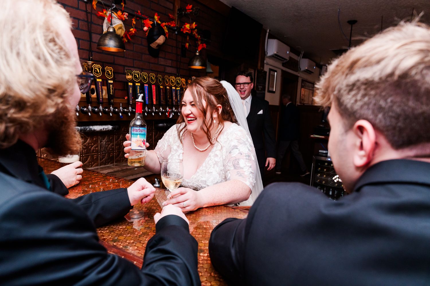 Wedding celebration scene at a rustic bar with guests raising glasses in a toast.