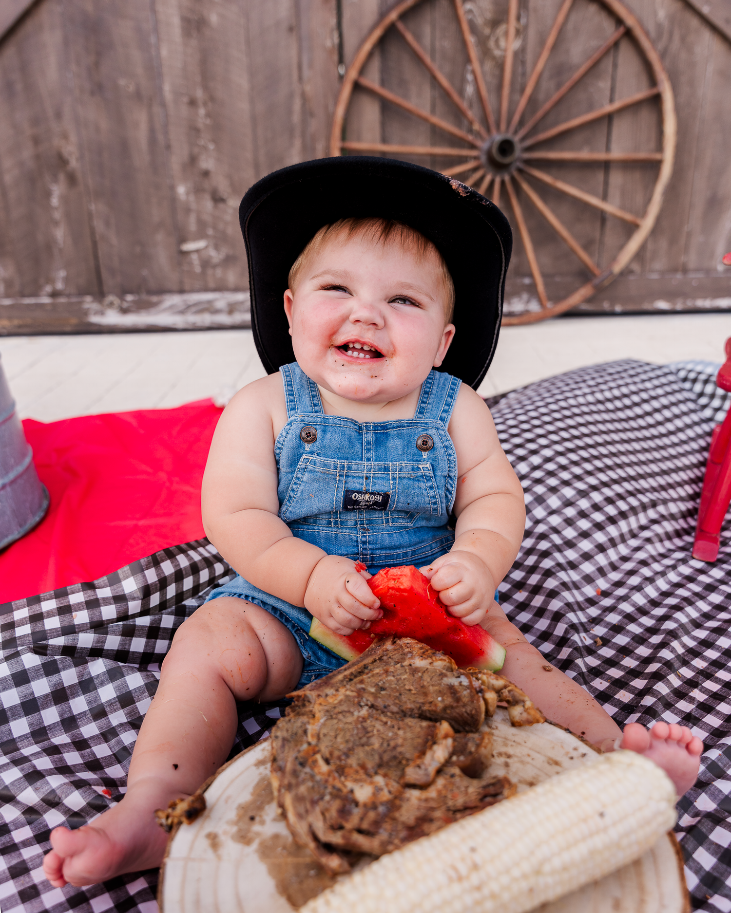 A baby in denim overalls sits on checkered pillows creating a mess while playing with bread.