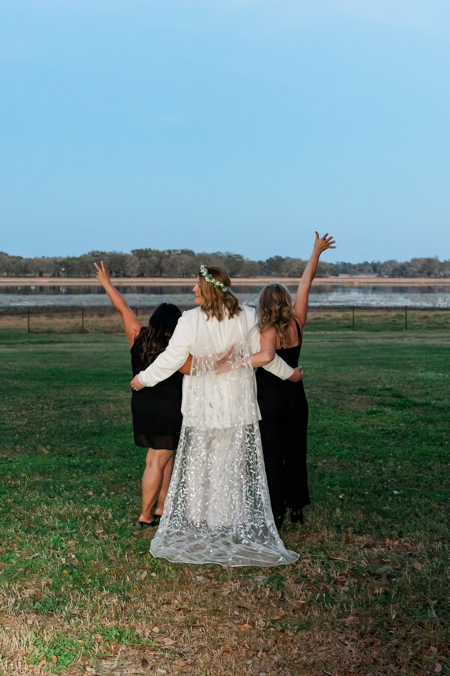 Three figures in formal wear raise their arms joyfully while standing on grass with a lake view in the background.