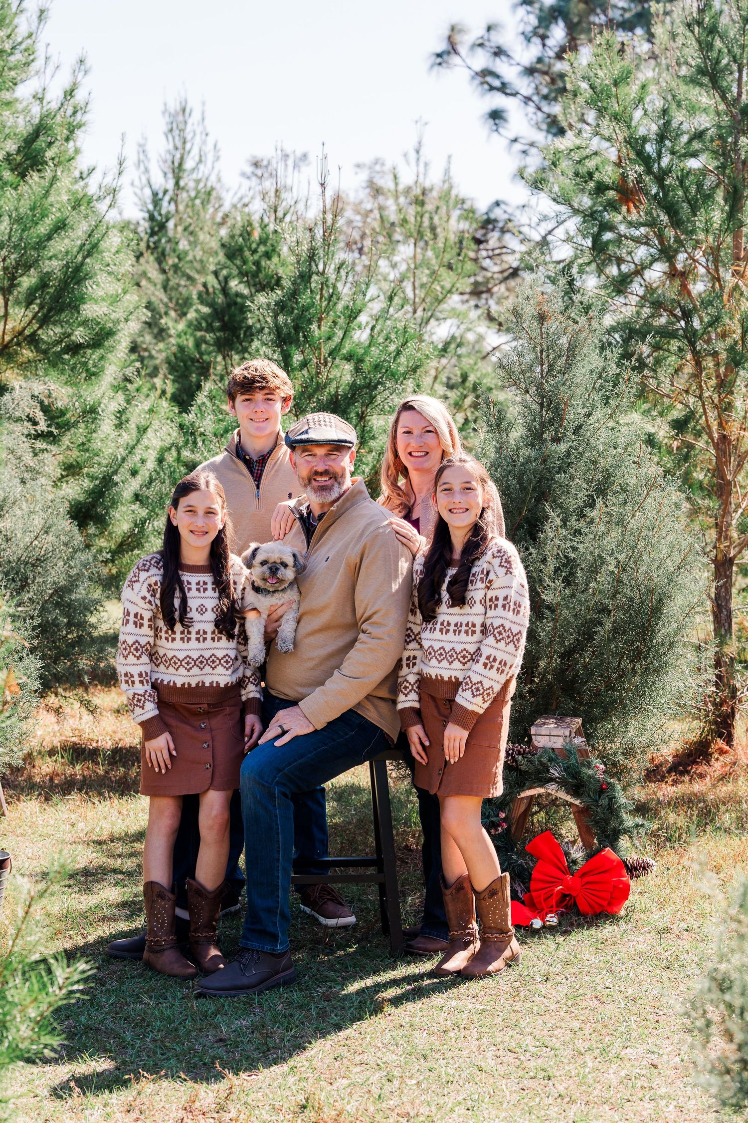 A family wearing coordinated brown and white winter sweaters poses together in front of evergreen trees outdoors.