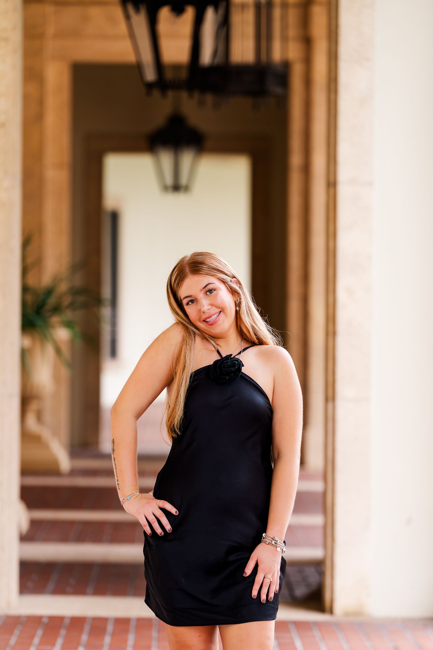 A woman in a sleeveless black dress poses in a covered outdoor corridor with columns and lanterns.
