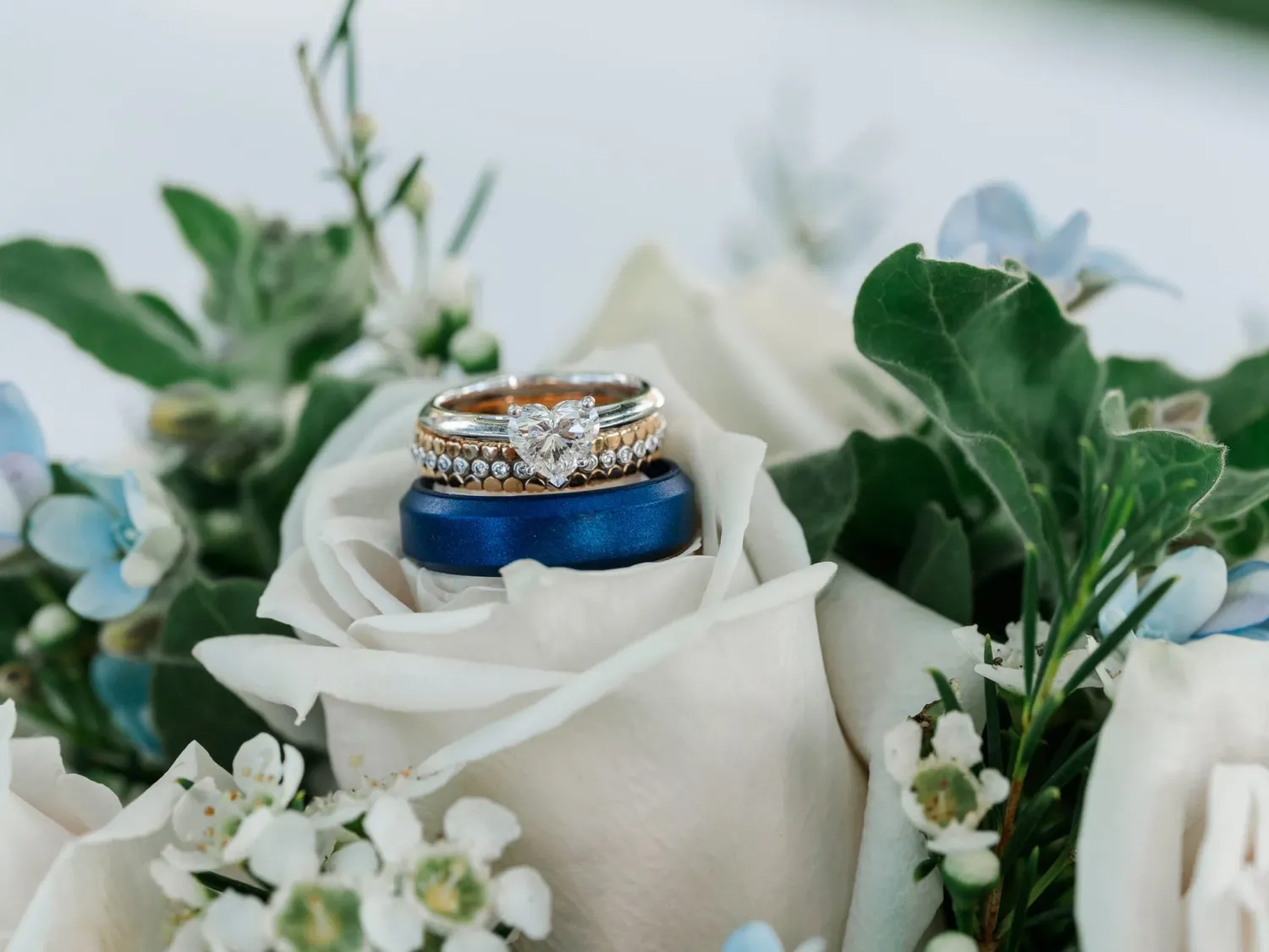 Wedding rings nestled on a white rose with blue accents and greenery in the background.