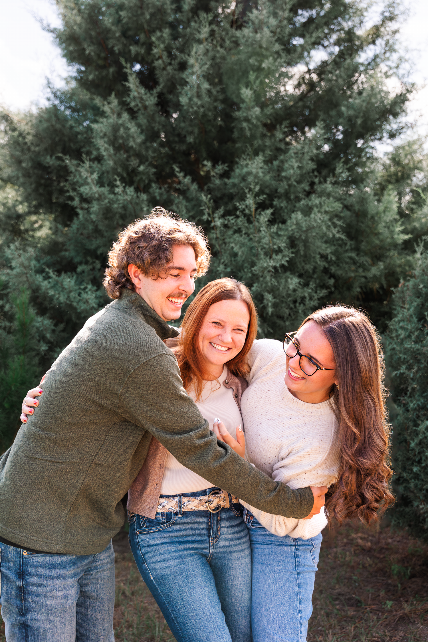 Three friends laughing and hugging outdoors near evergreen trees, wearing casual jeans and sweaters.