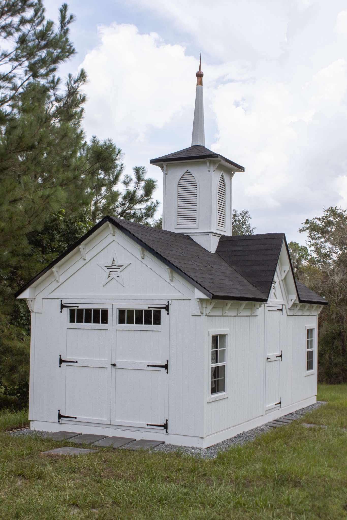 White miniature church building with steeple, dark roof, and barn-style doors sits on green lawn surrounded by trees.