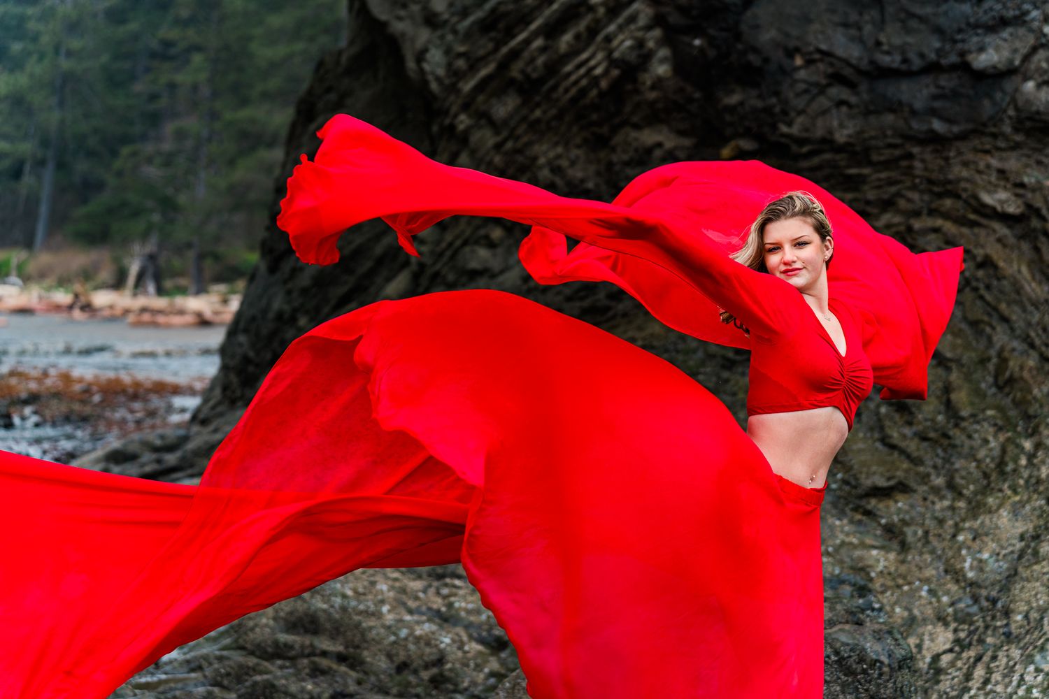 A flowing red dress billows dramatically against dark rocks near a shoreline.