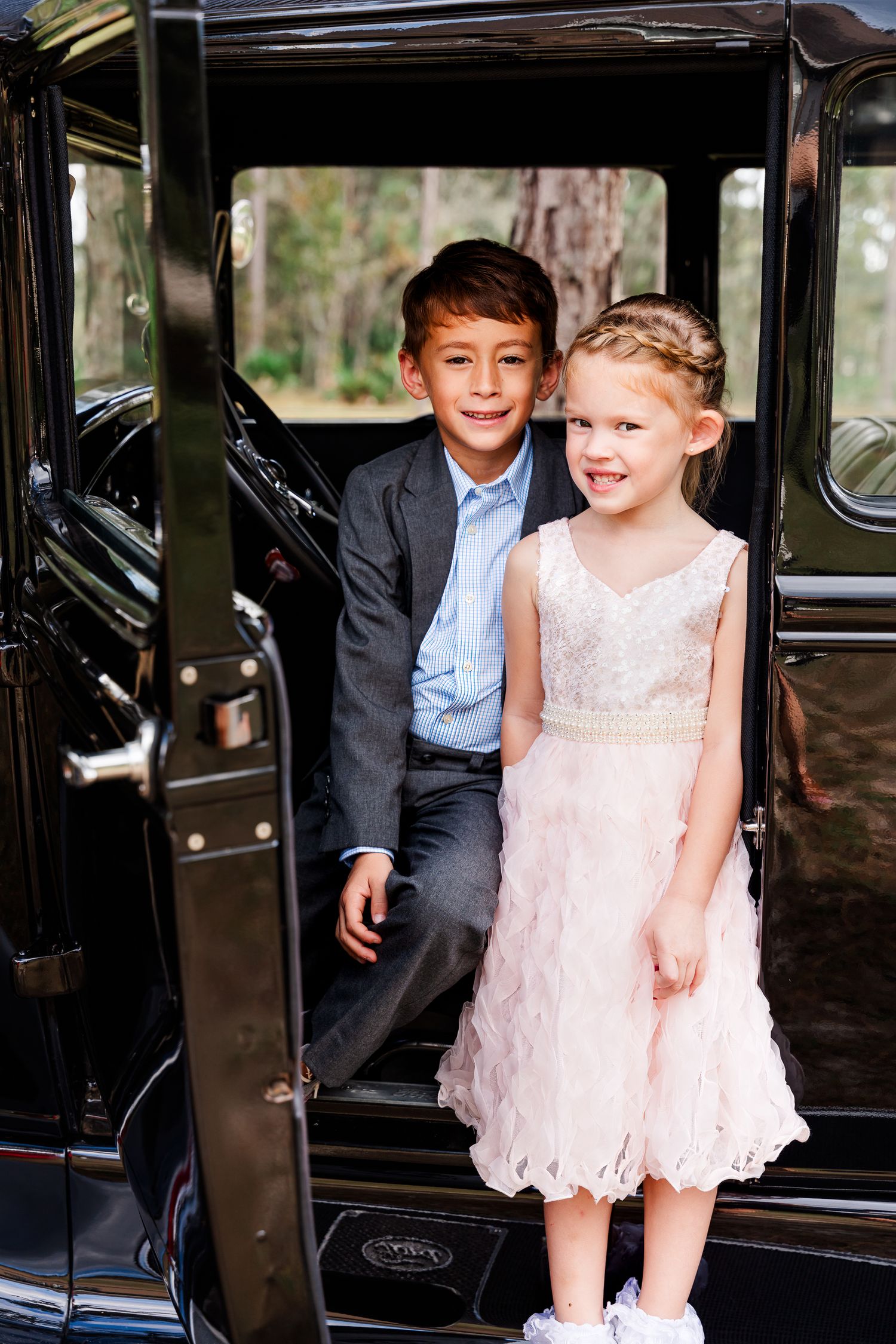 Two children in formal attire sitting in a vintage black car smiling at the camera.