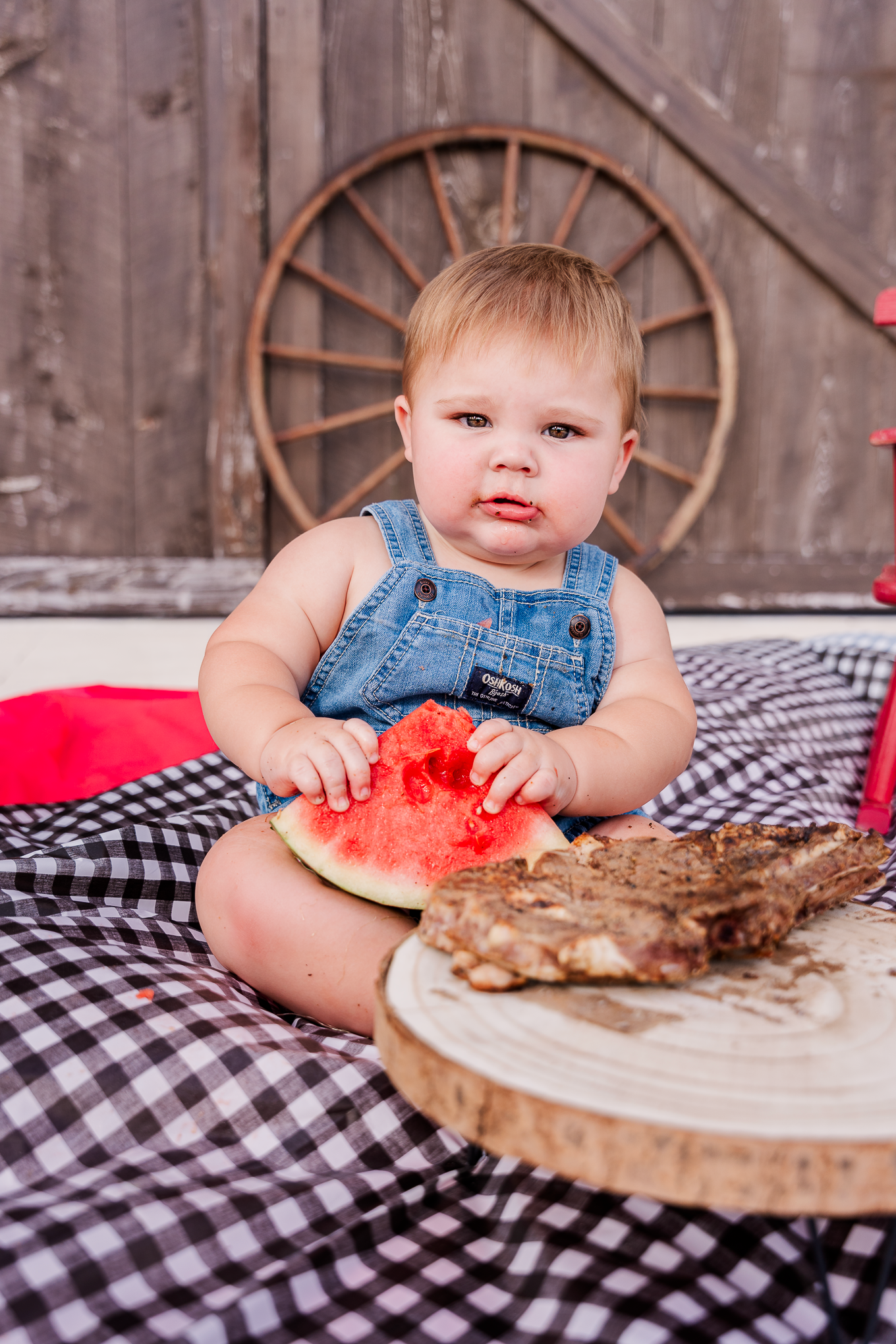 Toddler in denim overalls enjoys slice of watermelon on checkered picnic blanket with rustic wagon wheel backdrop.
